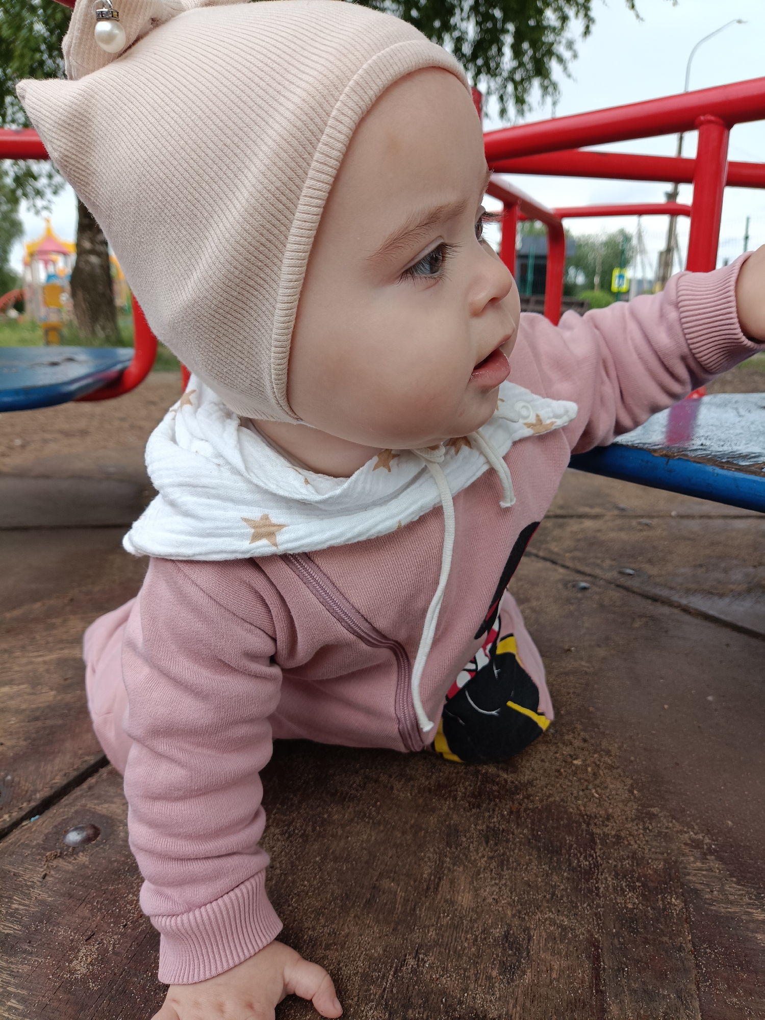 This image captures a baby on an outdoor playground in Chaikovsky, Russia, during what appears to be an overcast day. The baby, seen from a side profile, is positioned on a wooden platform covered with scattered dirt or sand, possibly in a crawling or leaning stance.
The baby wears a light beige ribbed knit hat, featuring a small silver ornament with a white pearl on its left side. A white scarf adorned with light brown star patterns is tied around the baby's neck. The main attire is a pink long-sleeved romper or onesie, which has ribbed cuffs and a partial character graphic in black, yellow, and red on the torso.
The baby's left hand is on the wooden platform, while the right arm reaches towards or rests on a blue metal bar of the playground equipment. A prominent red metal bar is visible in the mid-ground, extending horizontally. The baby's gaze is directed to the right, with a curious expression and a slightly open mouth.
The background is softly blurred, revealing green trees and distant colorful playground structures with yellow, blue, and red elements. A light pole and a yellow diamond-shaped road sign are also faintly visible, suggesting the playground's proximity to a public thoroughfare. No discernible text is visible in the image.