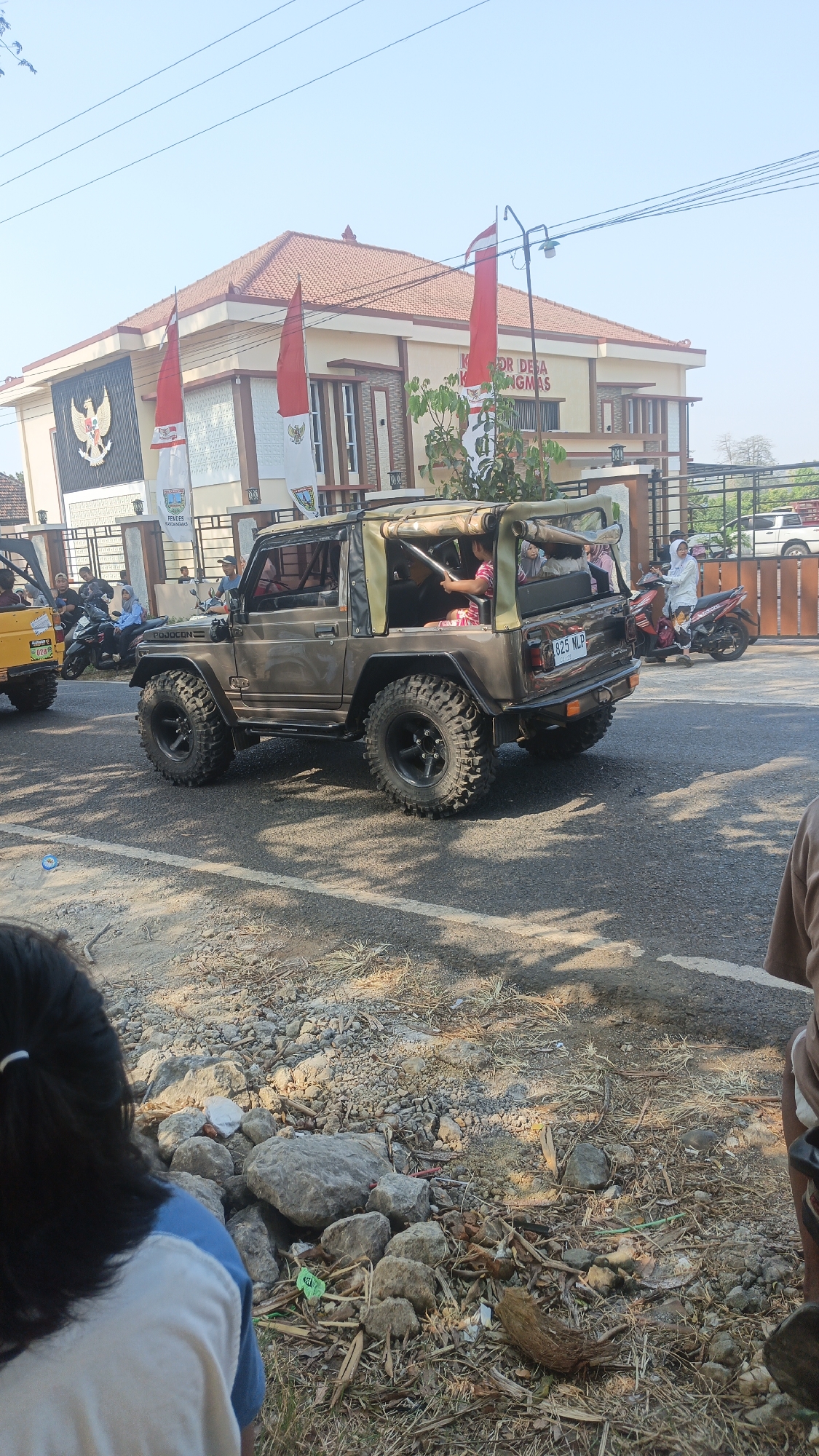 In the city of Kandangmas, Indonesia, under a clear sky, a convoy of vehicles moves along an asphalt road. The foreground is occupied by the back of a person's head and shoulders, their dark hair pulled back with a white clip. To the left, the rear of a yellow vehicle is visible. Dominating the mid-ground is a modified brown Jeep with large, off-road tires. The Jeep has its top down, revealing several occupants. In the background, a building with red-tiled roofs and cream-colored walls stands prominently, adorned with Indonesian flags and the national emblem. People on motorcycles can be seen on the right side of the road, with one individual in white riding a red scooter. The roadside is covered in gravel, dry grass, and scattered debris. The scene captures a moment of local activity on a bright day.