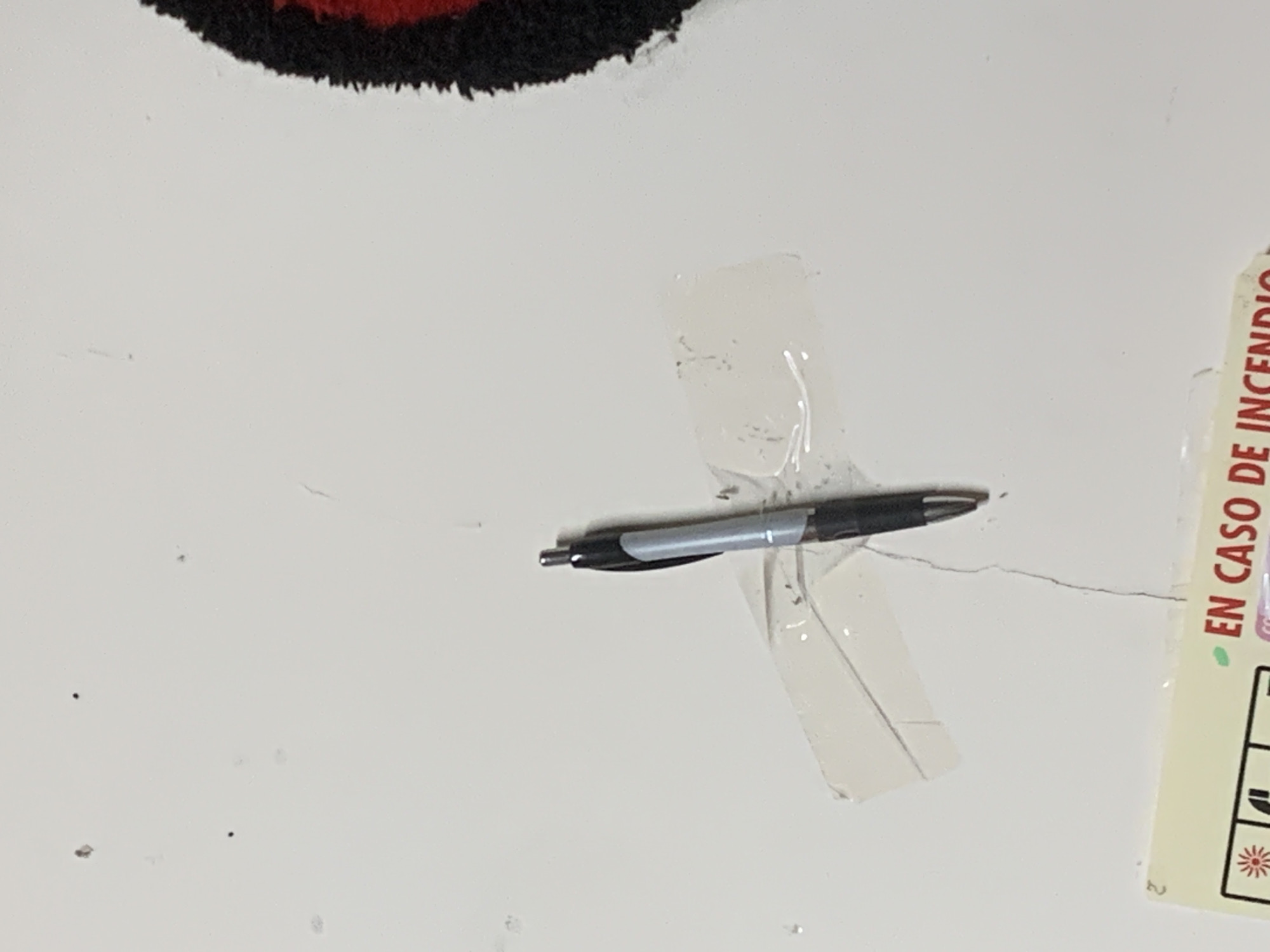 A black and silver pen is taped to a white surface with clear tape. Part of a rug is visible in the upper left corner, and a section of a label with the text "EN CASO DE INCENDIO" (In Case of Fire) is visible in the lower right corner. The location cannot be determined from the image.