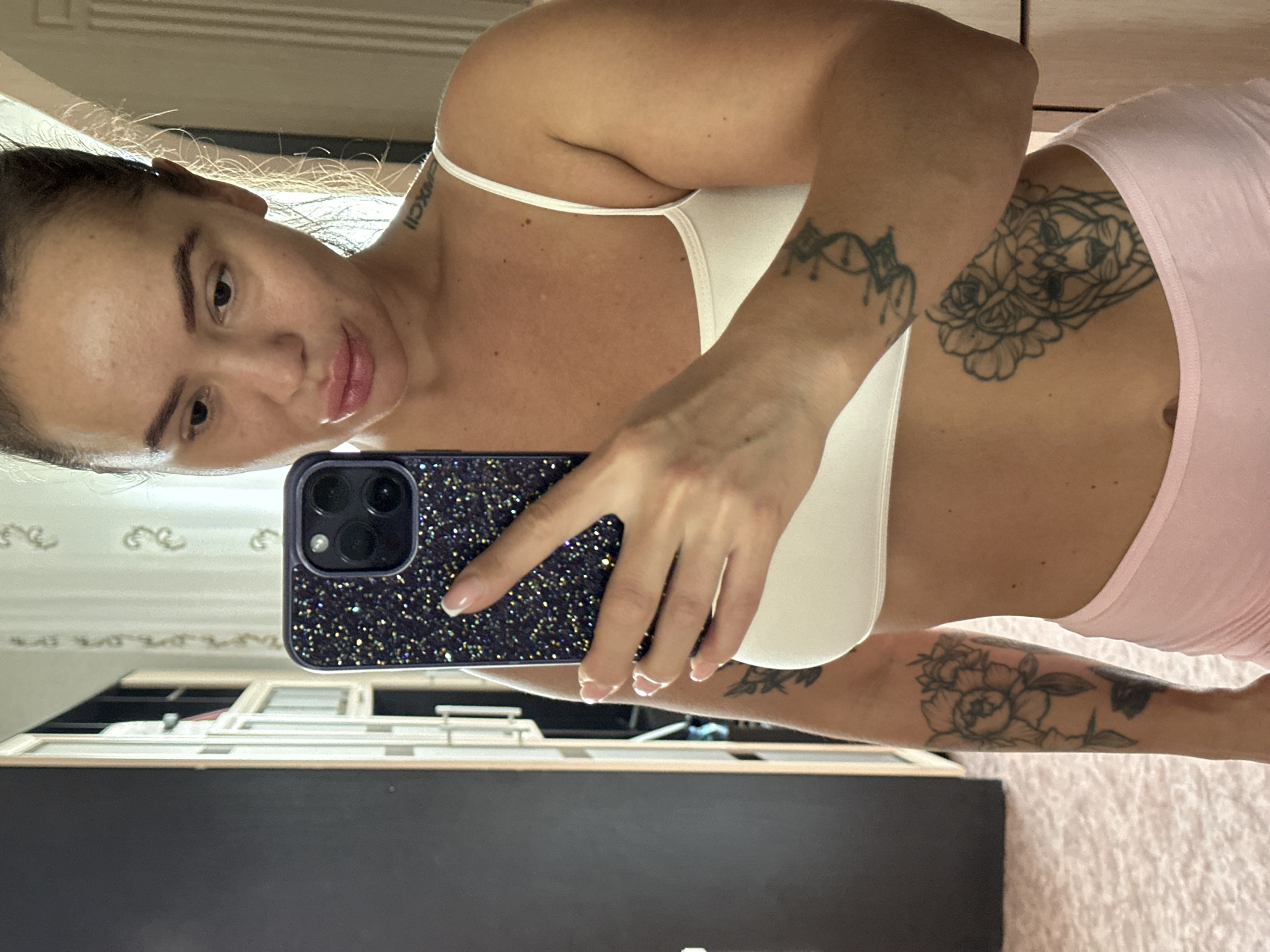 A medium shot captures a woman taking a mirror selfie in what appears to be a bedroom in Yuzhnouralsk, Russia. She is dressed in a white tank top and pink shorts, and her arms are adorned with tattoos. One tattoo on her right arm depicts a bouquet of flowers, while another on her left arm features a stylized lion's head crowned with a rose. She holds a dark, glitter-covered smartphone in her right hand, partially obscuring her torso. The background reveals a wardrobe and a wall with a subtle patterned wallpaper. The lighting suggests an indoor setting, likely during the daytime.
