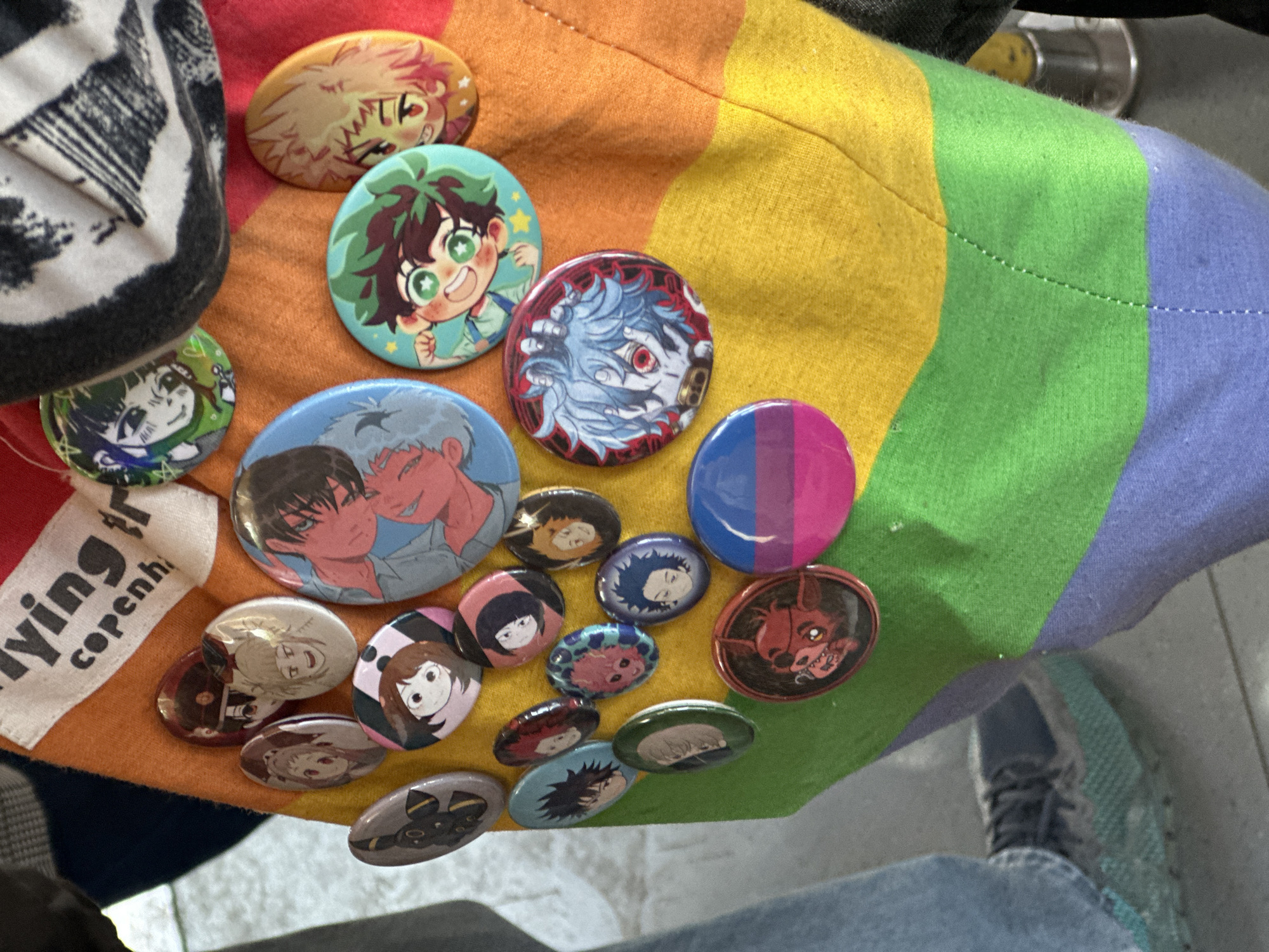 The image shows a collection of anime-themed buttons pinned to a rainbow-striped fabric, likely a bag or piece of clothing. The buttons feature characters from popular anime such as "My Hero Academia" and "Jujutsu Kaisen," alongside others including an Umbreon and Foxy from "Five Nights at Freddy's". There is also a button displaying the bisexual pride flag.
Other visible objects include part of a black and white patterned bag, a shoe, and a fragment of a garment (likely jeans).
Without further context, the location in Paris, France cannot be confirmed.