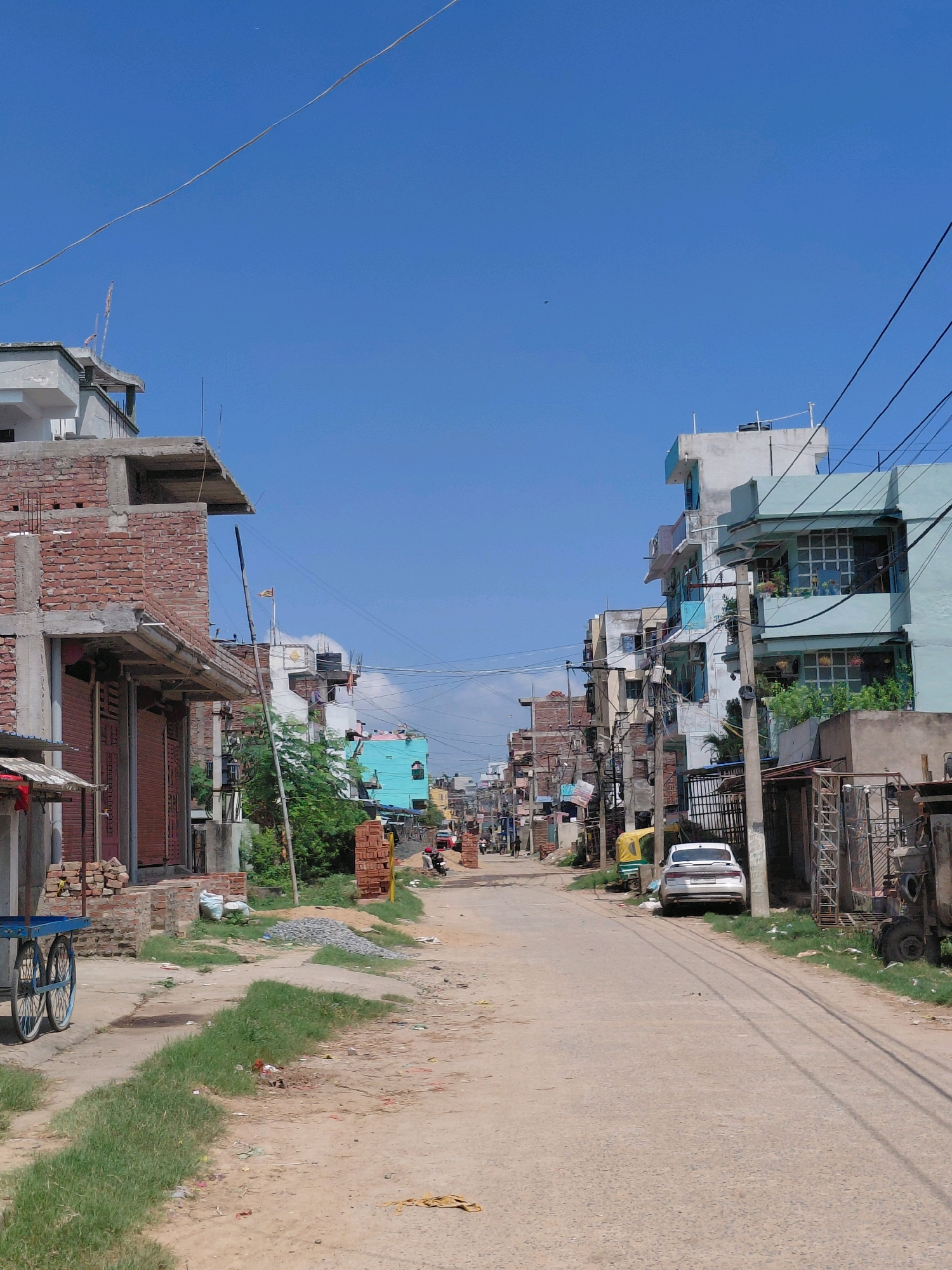 This is a street-level, eye-level, vertical, full-shot photograph of a dusty, unpaved road in Patna, India, likely in a developing urban area. The sky is a clear, bright blue, with a thin wire stretching diagonally across the upper portion of the frame. On either side of the road are multi-story buildings in various states of construction and completion.
On the left side of the road, in the foreground, is a partially constructed brick building with exposed brickwork and a concrete overhang. Metal shutters are visible on the ground floor, suggesting it might be a shop or commercial space. A blue cart with two large wheels and a wooden frame sits near the edge of the road in front of this building. Further down the left side, more buildings are visible, some painted in vibrant colors like turquoise, with a mix of balconies and windows.
On the right side of the road, buildings are also multi-story, with some painted in light blue and white. A white sedan car is parked on the side of the road. A yellow auto-rickshaw is visible behind the white car. Utility poles with a dense network of wires are prominent throughout the scene. Piles of bricks and construction materials are scattered along the sides of the road, indicating ongoing development. Patches of green grass grow unevenly along the road's edge, interspersed with dirt and debris. A few figures are visible in the distance, walking along the street, and a person is seated near a building on the right. The overall impression is one of a busy, but somewhat informal, urban environment undergoing growth and development.