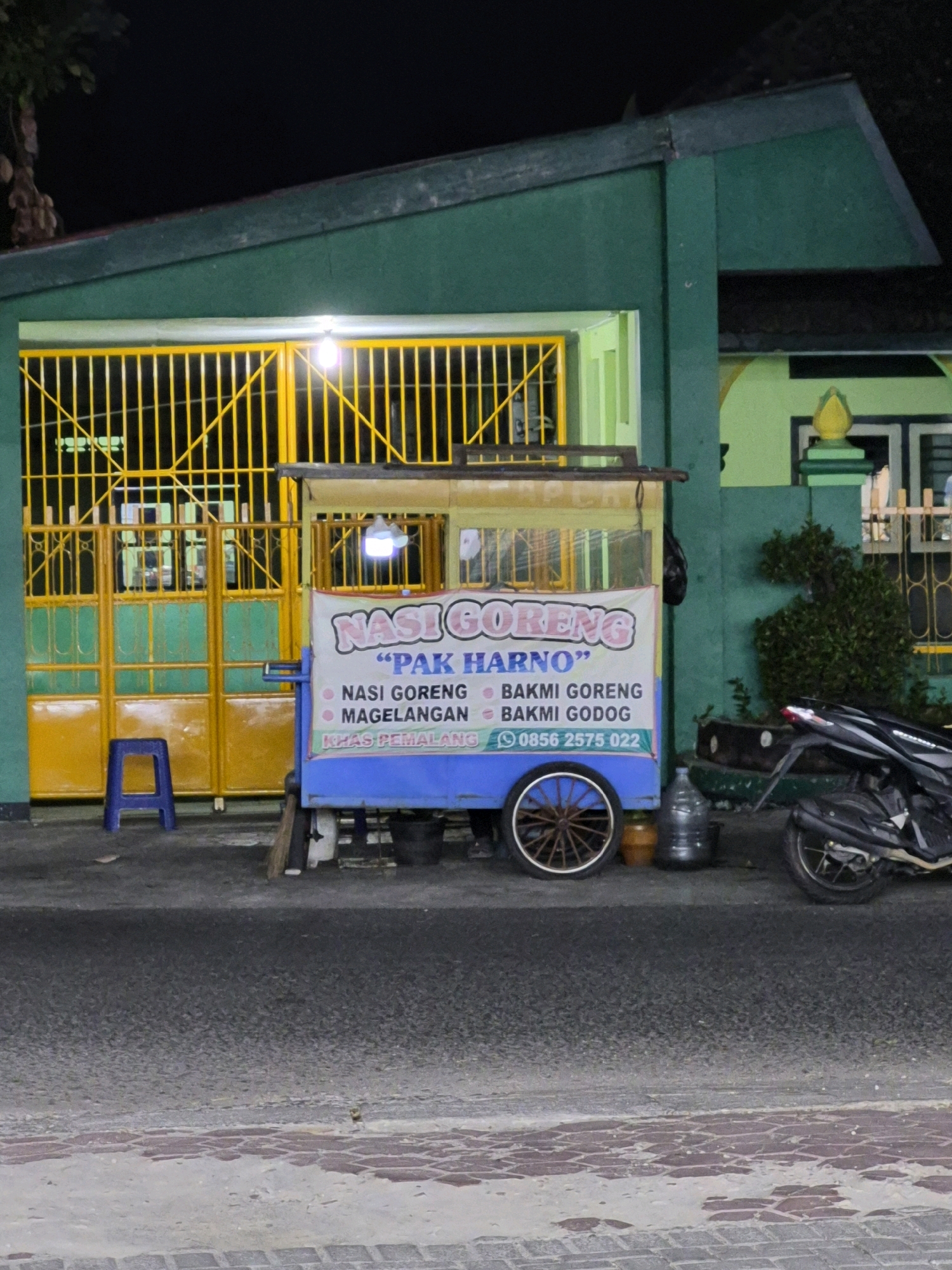 A food cart selling "Nasi Goreng Pak Harmo" is parked on the side of a street in Purworejo, Indonesia. The cart is blue and yellow and features a banner advertising its menu items: Nasi Goreng, Magelangan, Bakmi Goreng, and Bakmi Godog. A phone number, 0856 2575 022, is also displayed. A blue stool sits in front of a yellow gate, and a motorcycle is parked to the right of the cart. The building behind the cart is green with yellow trim. The street is paved with asphalt and bordered by brickwork. The scene appears to be set at night, with artificial lighting illuminating the cart and gate.