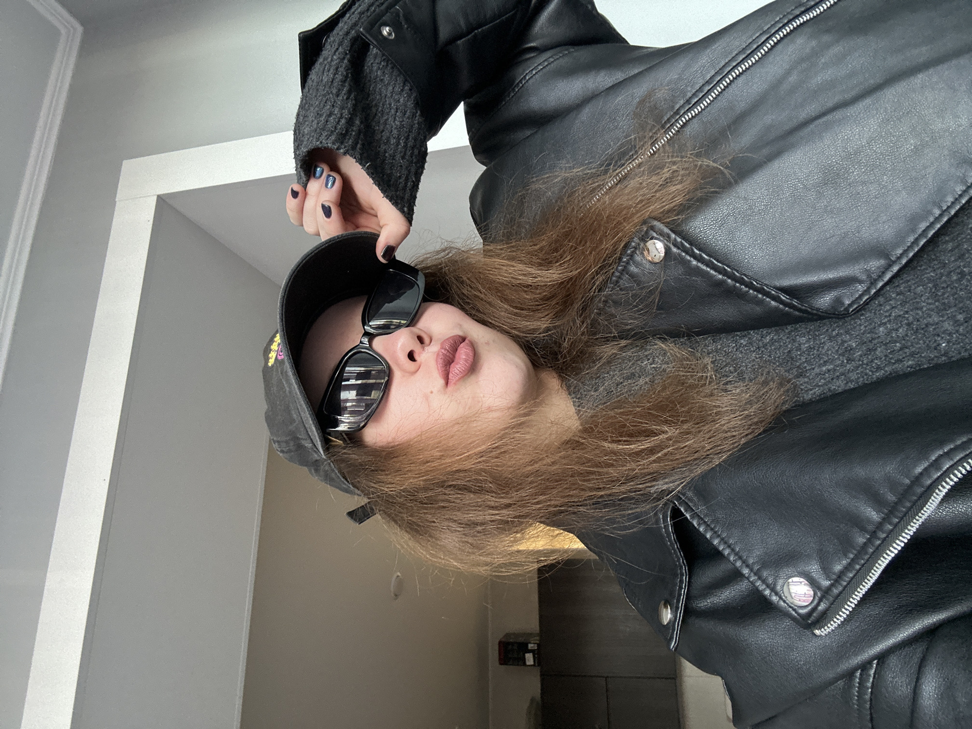 A woman with fair skin, long brown hair, and pink lips is positioned in the lower center of the frame. She is wearing a black baseball cap with an indiscernible design on the front and black-framed sunglasses with reflective lenses. She is wearing a dark gray sweater and a black leather jacket with silver zippers and buttons. Her left hand is raised, holding the brim of her hat, and her fingernails are painted in dark, iridescent shades of blue and purple. The background includes a white doorway in the upper left and a wall painted in shades of beige and gray in the lower right. There are objects and furniture faintly visible behind her.
The location in the city of Krasnoyarsk, Russia is unknown.