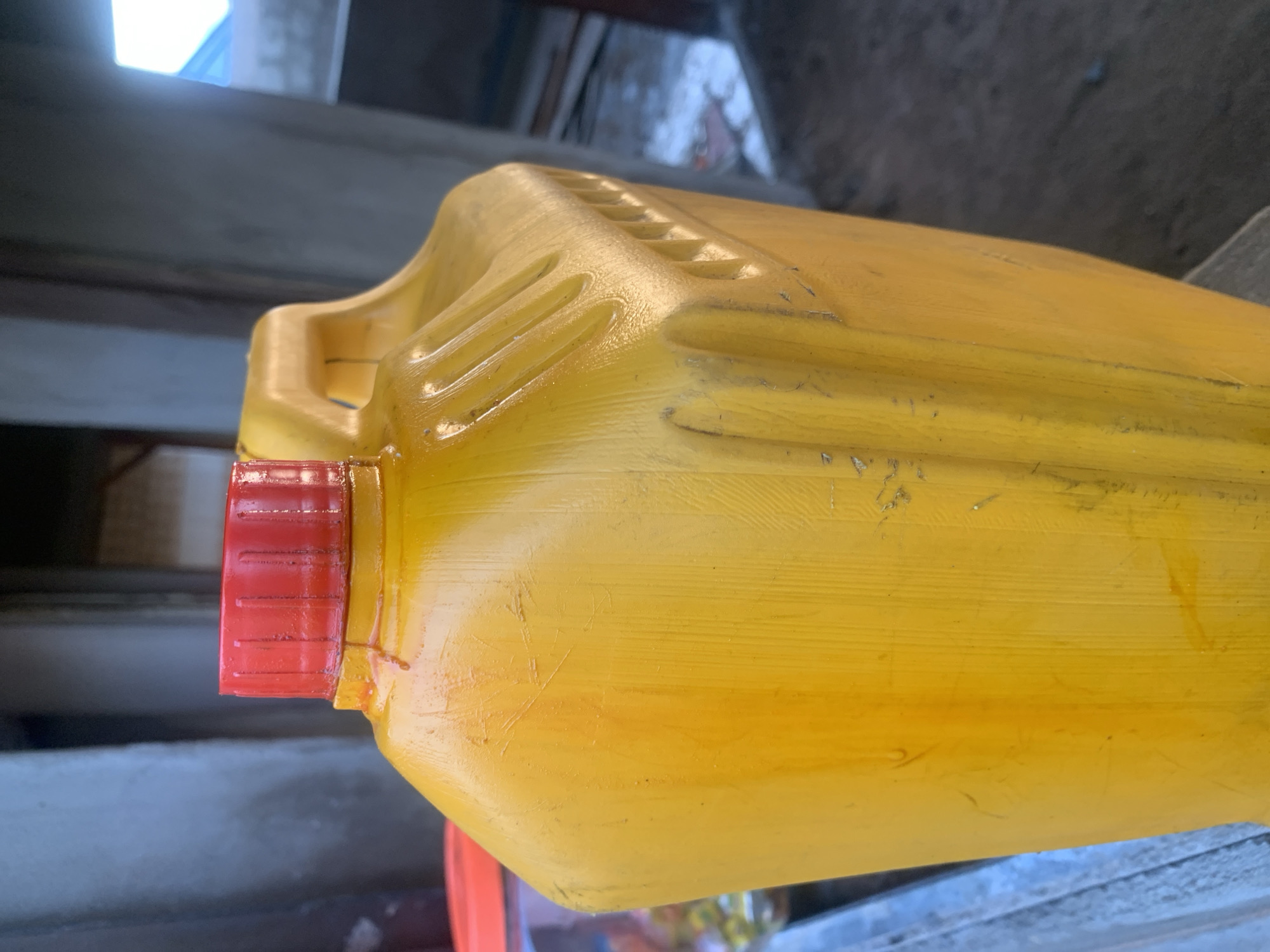 A close-up, eye-level shot focuses on a large, yellow plastic jug with a red cap, likely containing cooking oil or a similar liquid, placed outdoors in Jalingo, Nigeria. The jug is situated on a light blue, weathered surface, possibly a table or ledge. Behind the jug, the background is out of focus, but appears to be an outdoor setting with construction or unfinished structures. The jug has a textured surface with vertical ridges and visible scratches and smudges, indicating it is well-used. A handle is integrated into the top of the jug, and the red cap has a ribbed texture. The lighting suggests it is daytime, with a slight shadow cast on the surface in front of the jug. The overall impression is of a functional, everyday object in a utilitarian environment.