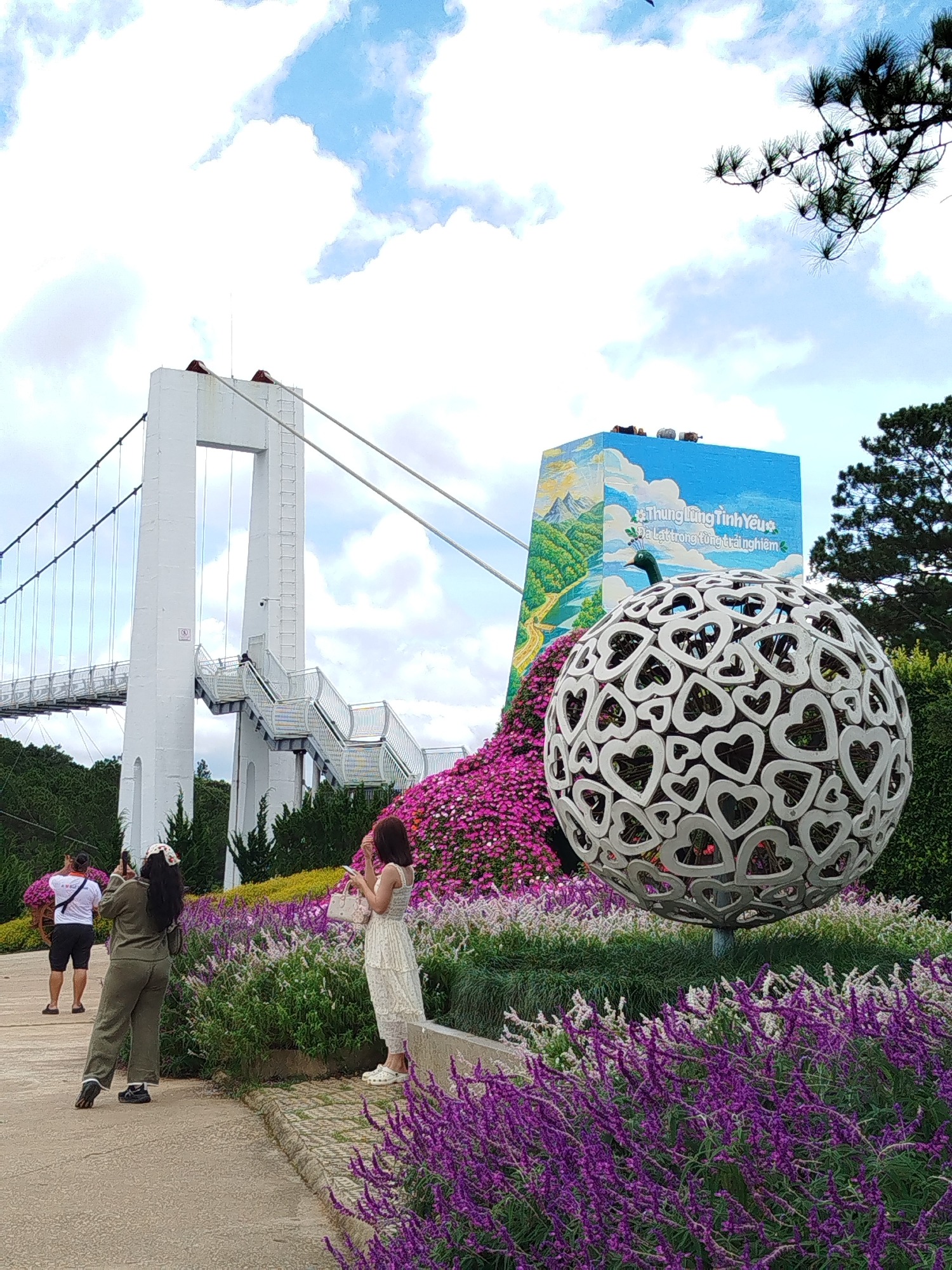This is a full-shot, eye-level, outdoor photograph taken in a garden setting, likely in Lâm Viên - Đà Lạt, Vietnam, given the signage in Vietnamese. In the foreground, there are vibrant purple and white flowers, some of which are in bloom. A large, spherical sculpture composed of interconnected heart shapes stands prominently in the mid-ground. Behind the sculpture is a colorful mural painted on a rectangular structure, depicting a landscape with mountains, a river, and a peacock. To the left, a suspension bridge with white towers and stairs is visible, partially obscured by trees. Several individuals are present in the scene; one person in a white shirt and black shorts is taking a photo with a phone. Another person, wearing a green jumpsuit and a patterned headband, is also photographing the scene. A third person, in a white dress and holding a purse, stands amidst the flowers, looking at their phone. The sky is cloudy with patches of blue.