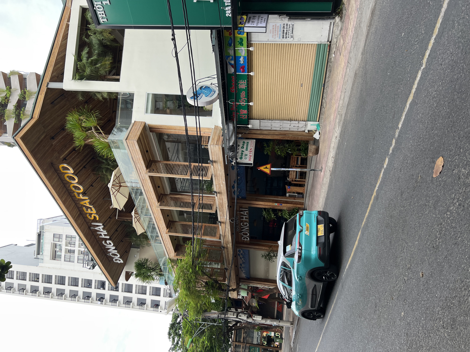 A bright teal and silver VinFast VF e34 electric car is parked on the right side of a paved street in Nha Trang, Vietnam. The car has a yellow license plate with black text and numbers. In the background, a multi-story building with wooden architectural elements and glass railings stands prominently. The building features large yellow lettering on its facade that reads "ĐÔNG HAI SEAFOOD." A tall, white building with many balconies is visible behind the seafood restaurant. A smaller shop with a closed roll-up door is situated to the right of the seafood restaurant, displaying Korean text and images of shoes. The street is lined with trees and power lines.