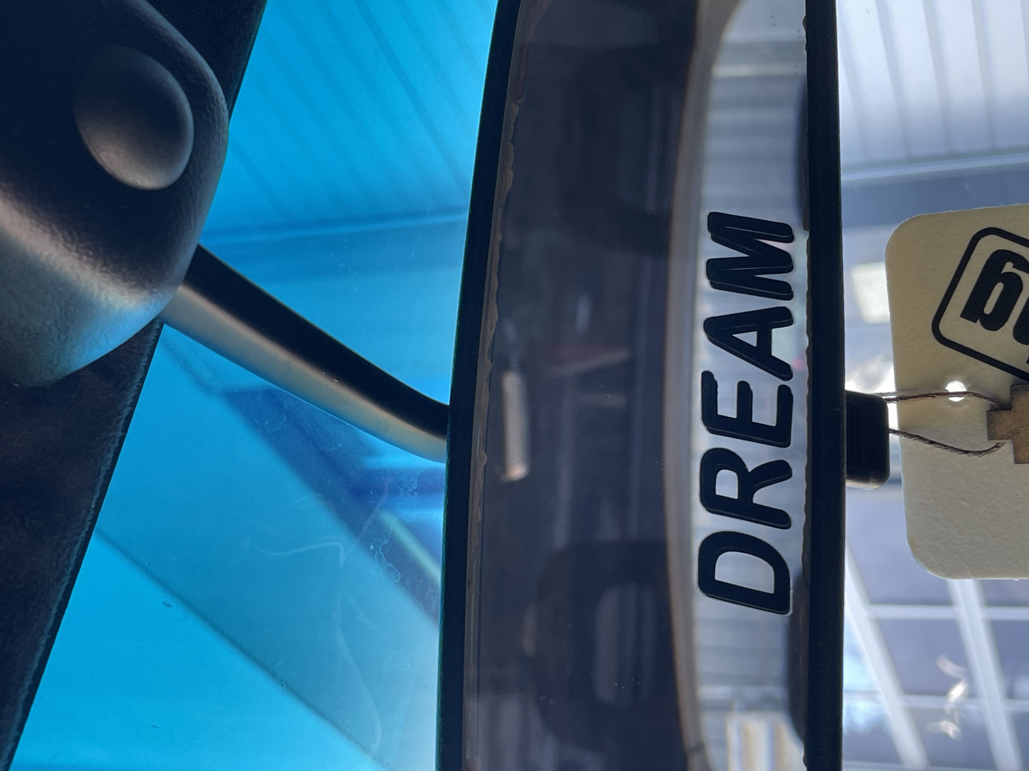 The image presents a close-up, obstructed view from inside a vehicle, likely a car, of a rearview mirror. The word "DREAM" is prominently displayed on the mirror's surface. A portion of a tag or card with the letter "b" in a square is visible, hanging from the mirror. The interior of the car is partially visible, with a blue-tinted element in the foreground. Through the windshield, a glimpse of an external environment is seen, likely an urban setting with building structures and a grid-like pattern suggesting windows. The location is listed as Unknown, Unknown.