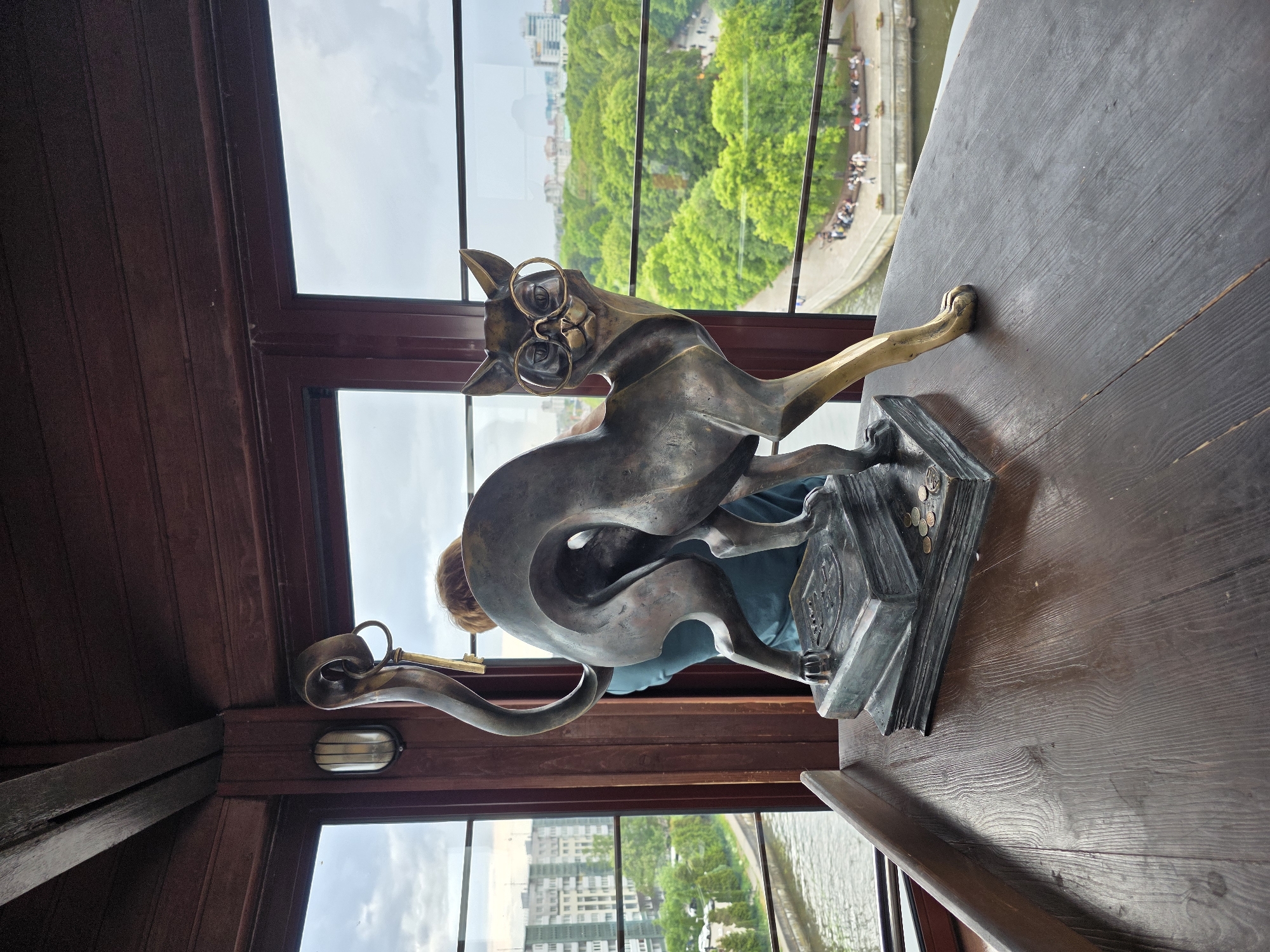 A bronze statue of a cat is prominently displayed on a dark, polished wooden table. The cat wears circular eyeglasses and stands on two stacked, dark bronze books. Several metallic coins are scattered on the top book at the base of the statue. A small golden-colored key hangs from the loop formed by the cat's upward-curled tail. The cat's front right leg is extended forward, exhibiting a lighter, more polished bronze finish compared to the darker, patinated body.
The interior setting features dark wooden paneling on the walls and ceiling, characteristic of a historical structure. Large windows frame a panoramic view of an urban landscape under a partly cloudy sky. A person with reddish-brown hair, wearing a teal-colored garment, is partially visible behind the statue, looking out the window. An oval, wall-mounted light fixture with a vertical bulb is affixed to the wooden window frame on the left.
The exterior view reveals a body of water, identifiable as the Pregolya River, bordered by a concrete embankment and lush green trees. Multiple groups of pedestrians are visible on the embankment and a paved walkway alongside the river. Multi-story residential buildings with light-colored facades are discernible on the left bank, with additional city structures in the distant background. This scene is located within the King's Tower (Koenigsturm) on Kant Island, part of the Museum of the World Ocean, in Kaliningrad, Russia.