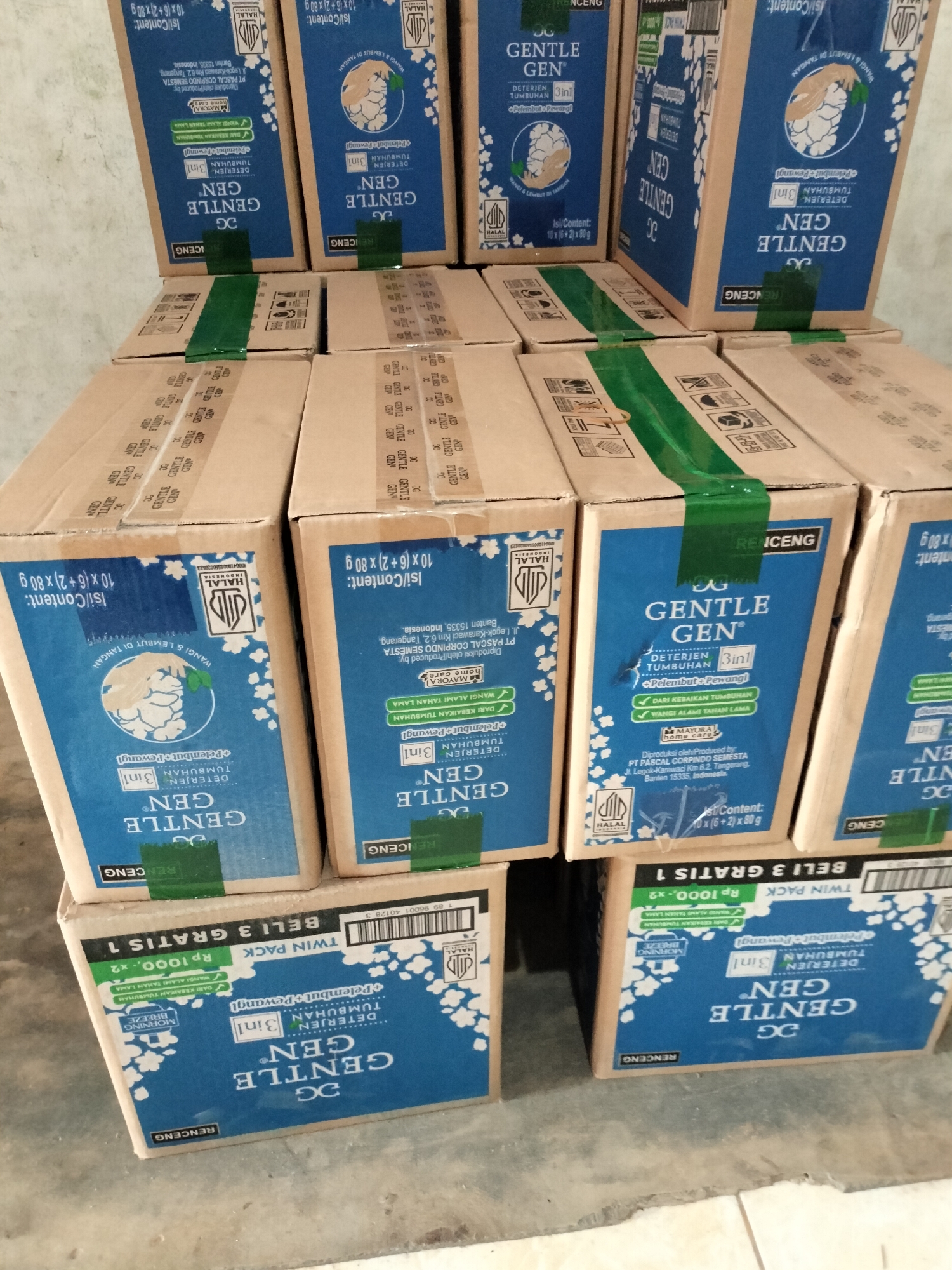 The image shows a stack of cardboard boxes with blue and white labels, stacked on a gray concrete floor. The boxes appear to contain laundry detergent. Some boxes are upright, while others are on their sides. Green tape is used to seal some of the boxes. The labels on the boxes feature the brand name "GENTLE GEN" in large, white lettering. Text on the labels, in Indonesian, indicates the product is a detergent with a fabric softener and fragrance, and it is suitable for plant-based cleaning. One of the boxes has text that translates to "Buy 3 Get 1 Free" and "Twin Pack Rp 10000.-x2". The overall impression is one of commercial packaging, likely for retail or storage. The image does not contain any specific landmarks or contextual clues to indicate its location in Langsa, Indonesia, or any other city.
