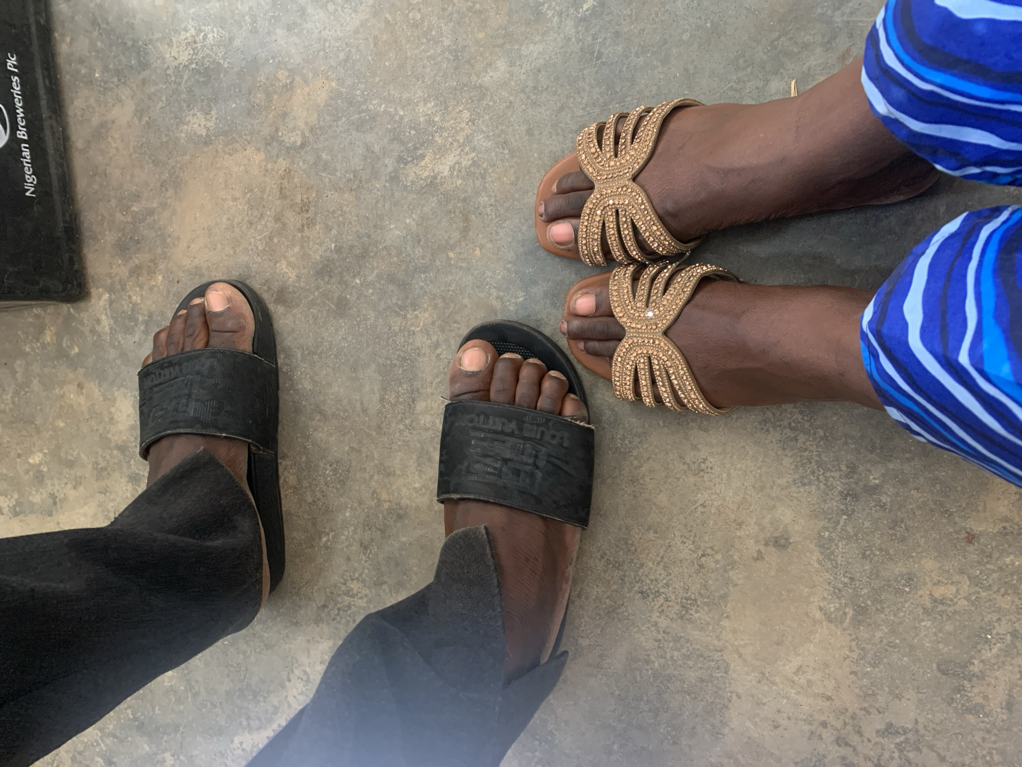 The image depicts two pairs of feet on a gray concrete surface. The feet on the left wear black slide sandals with the inscription "LOUIS VUITTON". The feet are clad in black pants. The feet on the right are in tan sandals with a jeweled, decorative design. These feet are next to blue and white striped fabric. A black object with the partial inscription "Nigerian Breweries Plc" is partially visible in the upper left corner of the image. The scene likely takes place in Kabong, Nigeria, although there is no direct confirmation beyond the inscription.