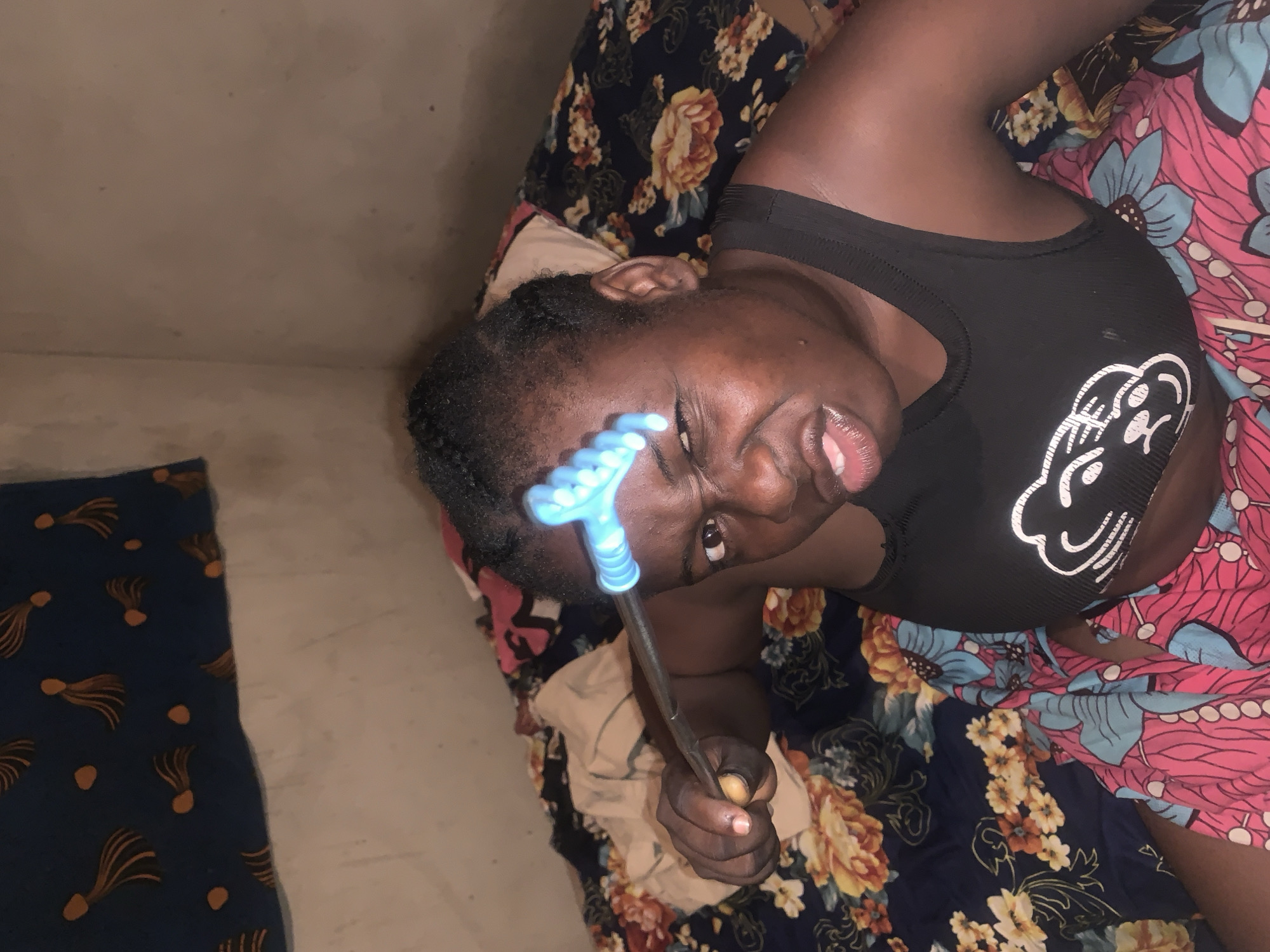 A woman lies on a bed with floral-patterned sheets in what appears to be a room in Agban, Nigeria. She is holding a blue, rake-like tool to her forehead. She is wearing a black tank top with a white graphic. The room has a light-colored wall. A piece of dark blue patterned cloth is on the floor.