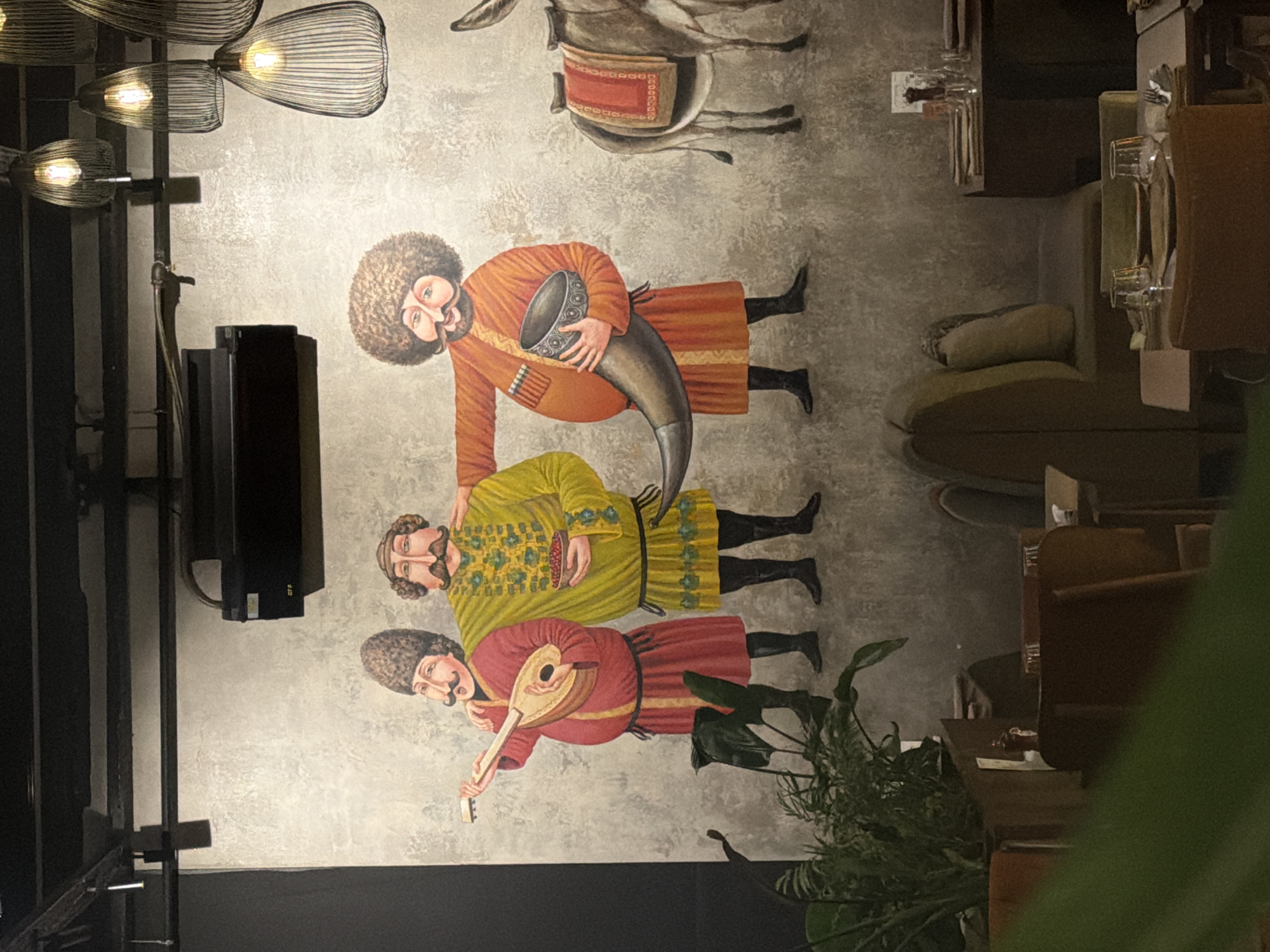 The image shows a mural painted on a textured grey wall inside a restaurant. The mural depicts three men in traditional attire and a donkey on the right. The leftmost man, wearing a red robe and a fur hat, plays a lute-like instrument. The middle man, in a yellow robe adorned with green and gold embroidery, holds a bowl of red berries. The rightmost man, in an orange robe and a large curly wig, holds a large horn in his right hand. The donkey is saddled with a red blanket. In the foreground, out of focus, are tables with settings, chairs, and lush green plants. The restaurant ceiling has exposed metal beams and hanging light fixtures. The overall scene is likely from a restaurant in Kadıköy, Turkey, given the style of art which may represent local or regional culture.