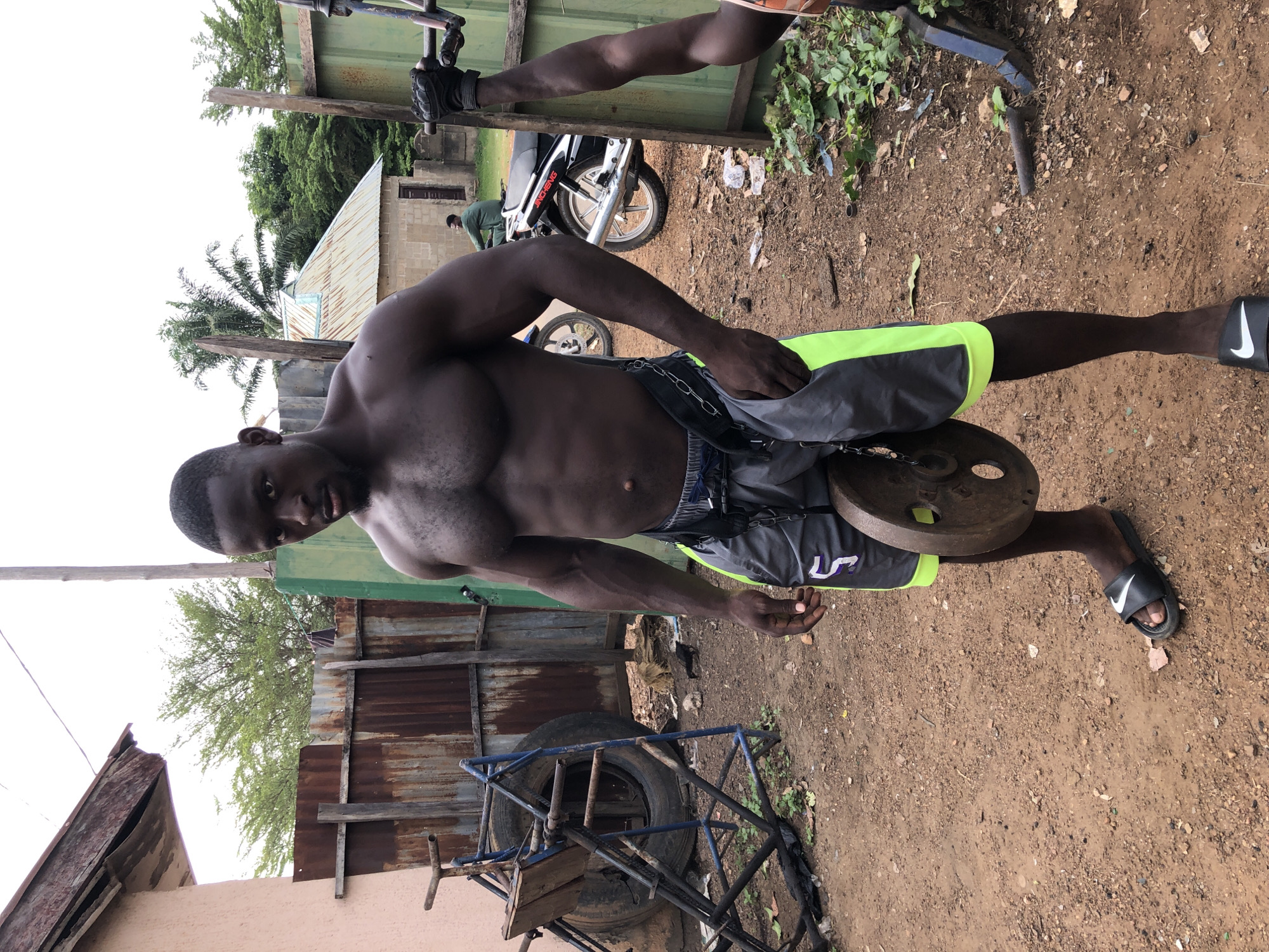 A shirtless Black man is centered in the frame, standing on a dirt ground in what appears to be an outdoor, makeshift gym setting. He is wearing grey and neon green shorts and black Nike slides. A metal weight plate is suspended between his legs by a chain.
In the background, partially obscured by the man and various pieces of equipment, there are tin structures. The presence of a motorcycle and another person in the distance suggests an inhabited area. The scene is cluttered with various pieces of metal, tires, and other materials used for building exercise equipment.
The location is Akwanga, Nigeria.