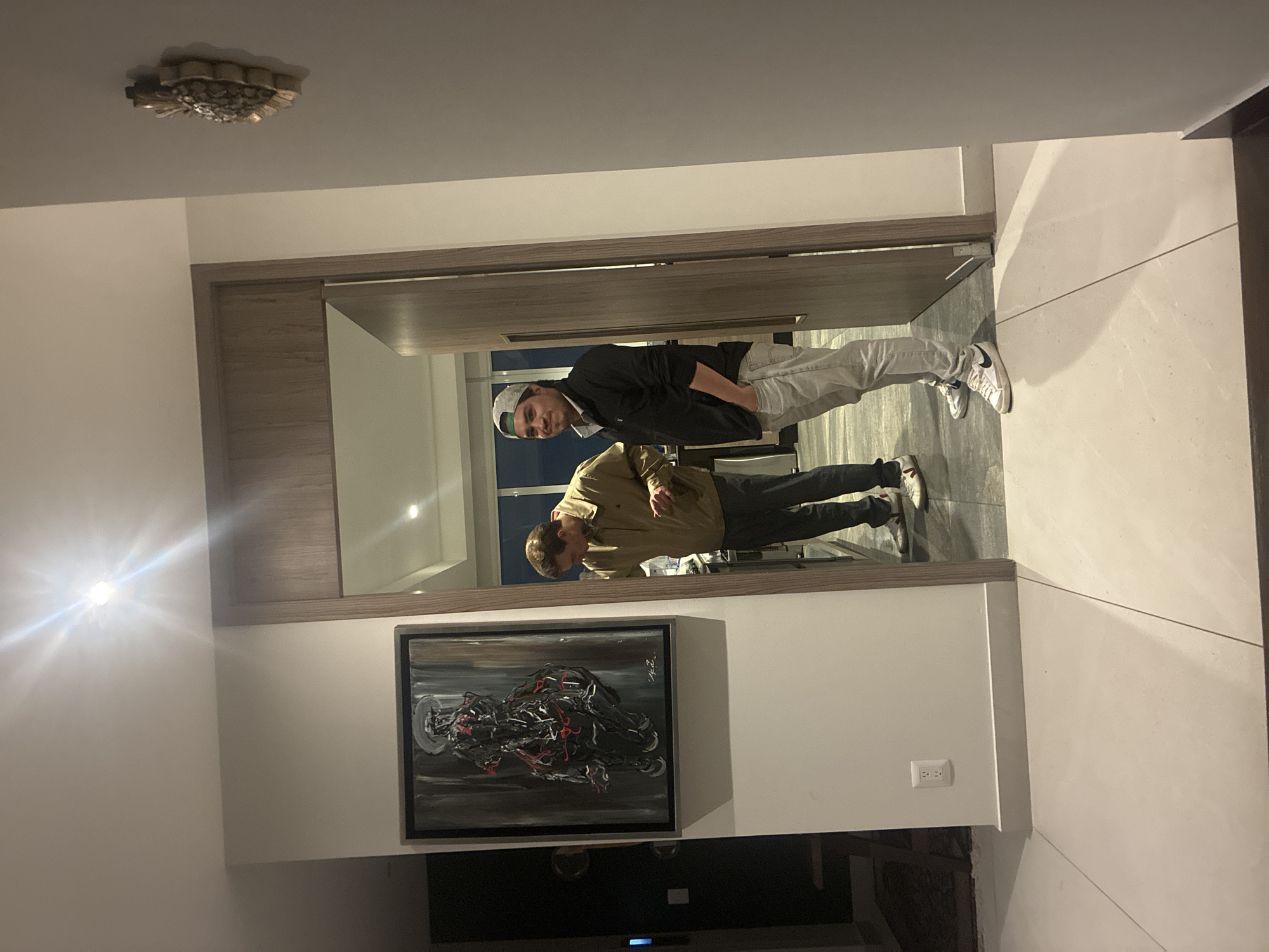 Two males are reflected in a large mirror in an indoor hallway. The hallway has white walls, a dark stone tile floor, and recessed lighting. The mirror is framed with dark wood and is mounted on a wall above a piece of abstract art. An outlet is visible on the wall beside the painting. In the reflection, the man closest to the mirror is wearing a tan jacket and dark pants. The other man is wearing a dark jacket, grey pants, and a baseball cap. A decorative ceiling light fixture is visible in the upper left corner of the image. The location is Huixquilucan de Degollado, Mexico.