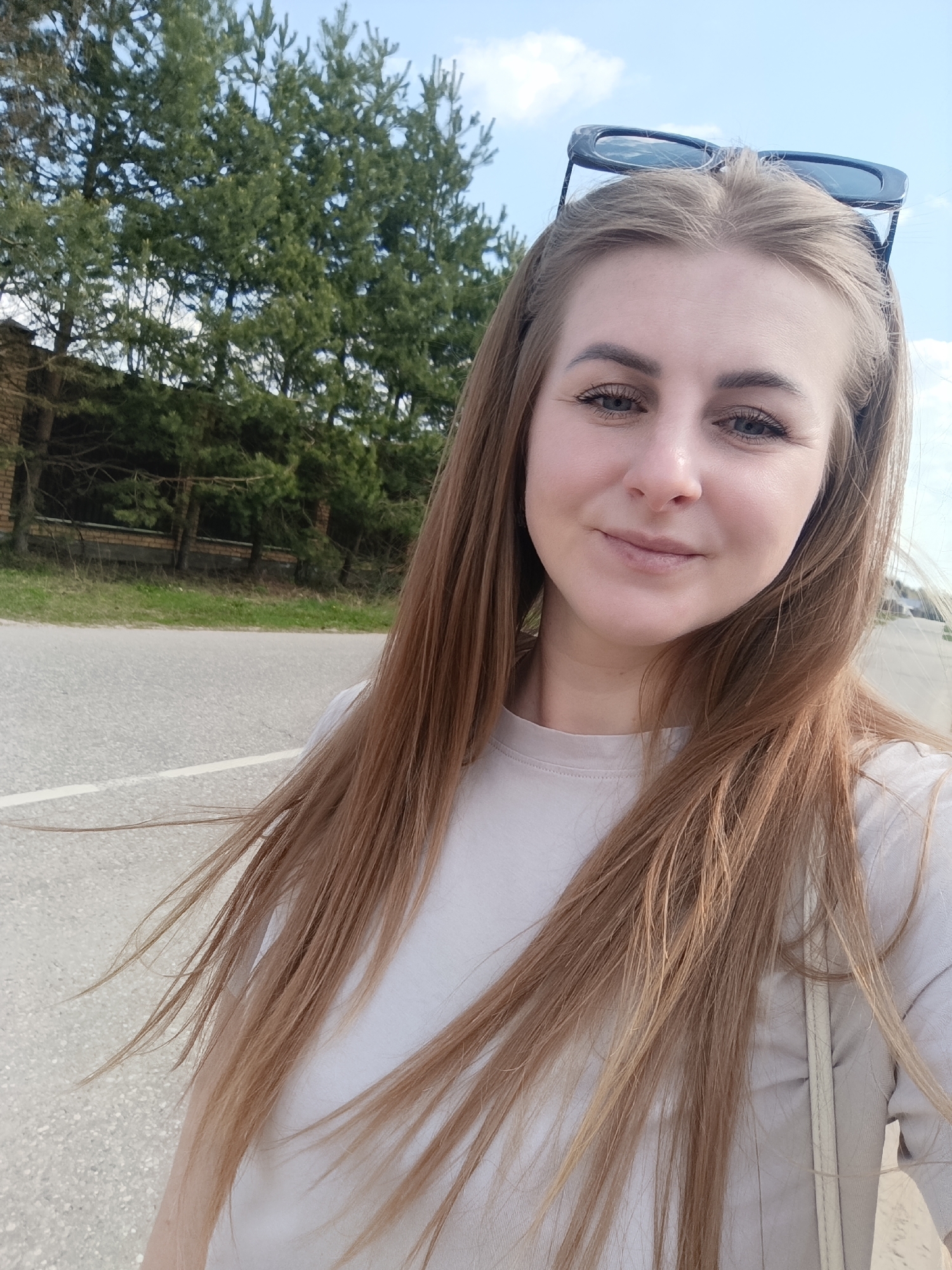 A woman is standing on a road on a bright day. She has long, light brown hair, sunglasses perched on her head, and is wearing a light-colored t-shirt. The road is asphalt and has a white line painted on it. Behind the woman and the road is a brick wall and some green trees. The sky is blue with a few clouds. The picture appears to be taken in the city of Ivanovskoe, Russia.