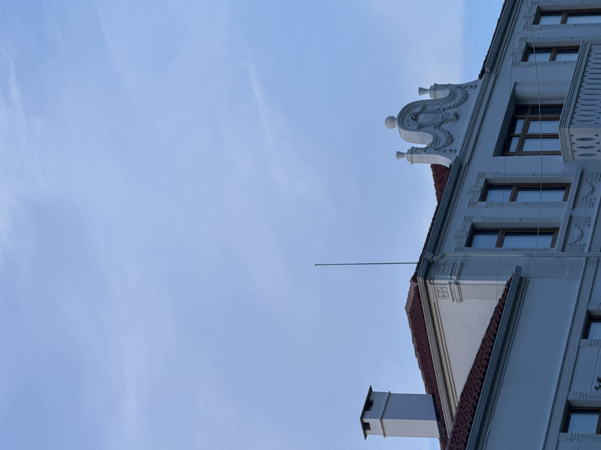 This image showcases the architectural details of an ornate building against a pale blue sky with wispy clouds, suggesting a clear day. The building, likely in Sozopol, Bulgaria, features decorative elements like elaborate stucco work on the facade and a red tiled roof. Numerous windows with dark frames are visible, suggesting multiple stories. A metal rod extends from the building into the sky, possibly an antenna or lightning rod. A chimney made of light-colored bricks and a white rectangular structure, possibly part of the building's ventilation system, are also present. The lighting indicates it is either early morning or late afternoon. The image is composed from a low angle, emphasizing the building's height and the vastness of the sky. No people or explicit activity are depicted, focusing instead on the building's static grandeur and the atmospheric conditions.