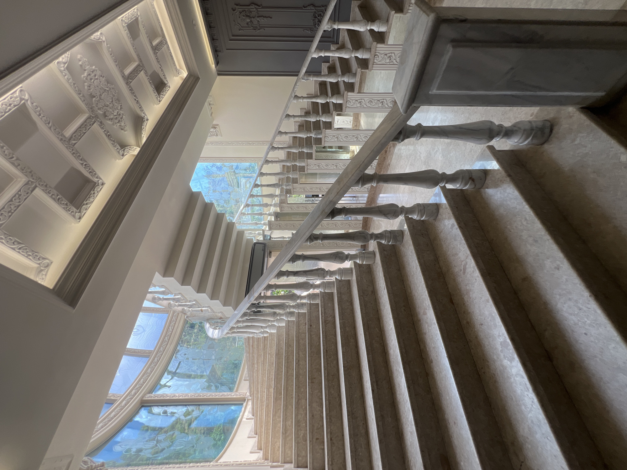 An interior shot of a staircase in Yerevan, Armenia, looking downward. The staircase is made of marble with a marble handrail and decorative balusters. A large window with blue stained glass provides natural light. The ceiling is ornate with detailed plasterwork. A closed door is visible at the top of the stairs. No people are visible.