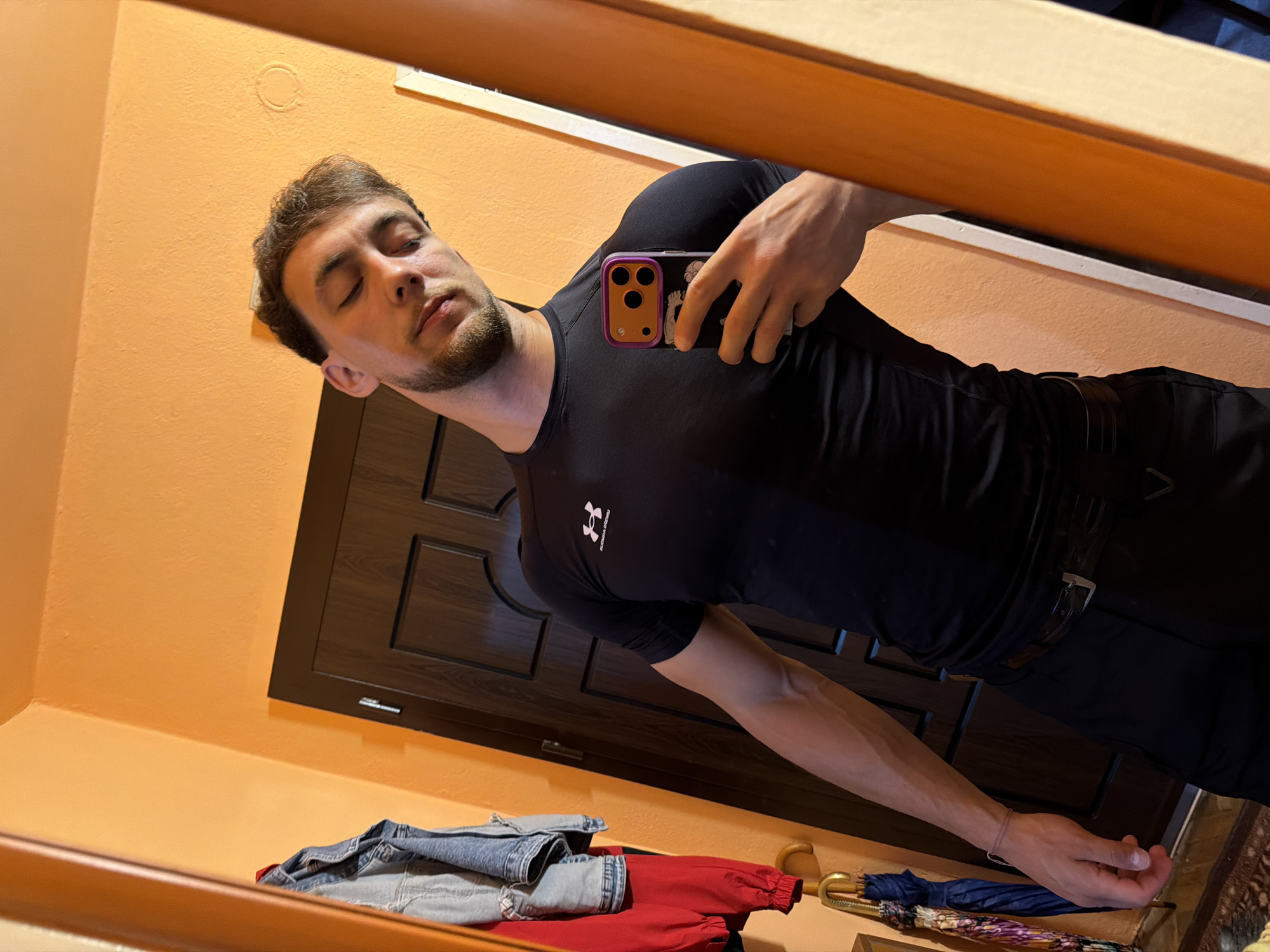 This image depicts an indoor mirror selfie taken in Tsarevo, Bulgaria. The subject, a male with light skin, short brown hair, and a short beard, is positioned facing forward, capturing his reflection. His eyes are closed, and his head is tilted towards his left shoulder. He wears a form-fitting black short-sleeved compression shirt, featuring a white Under Armour logo on the chest with the text "UNDER ARMOUR HECTIC" below it. He also wears black trousers secured by a black belt with a silver buckle. His right hand holds a dark-colored iPhone, equipped with a distinct purple and orange protective case that displays a visible triple-lens camera system and a stylized white wave graphic on its back. A thin, light brown string wristband is present on his left wrist.
The background, as reflected in the mirror, consists primarily of an orange-colored wall. A dark brown wooden door with paneling and a silver handle is visible behind the subject. To the far left of the frame, a wooden coat rack or hanger stand supports a light blue denim jacket, a folded red garment, and a blue umbrella with a distinct pattern hanging beneath them. The lower portion of the scene reveals a patterned floor surface, likely a rug or carpet. The mirror itself has a visible wooden frame on the right side.