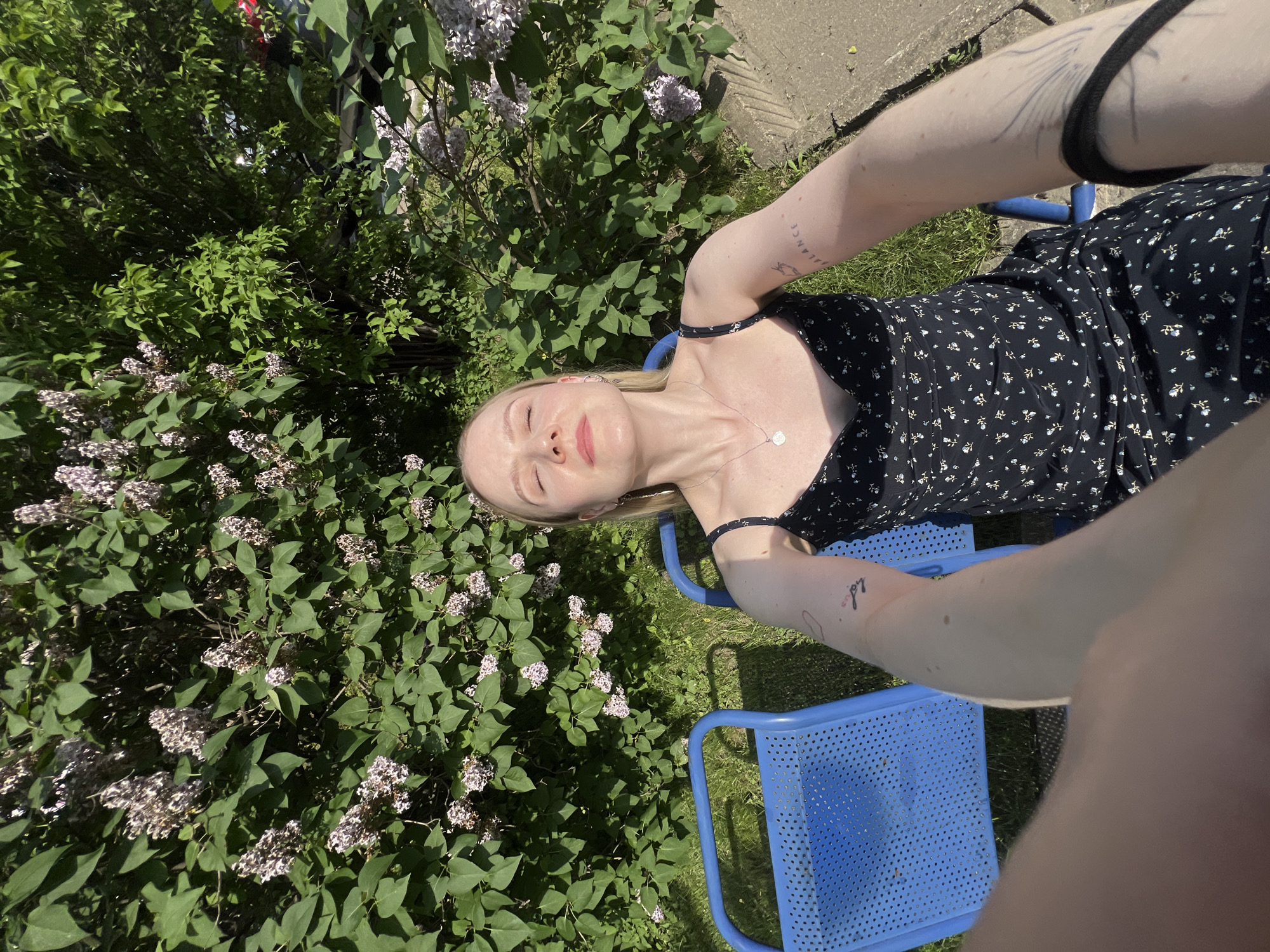 A young fair-skinned woman is reclining in a blue metal lawn chair outdoors. She wears a black floral dress and a silver necklace. She has tattoos on her arms. Lilac bushes with blooms are on the left side of the frame. The ground appears to be grassy with a concrete slab visible in the background. The image is taken at an unusual angle, appearing as a selfie. The location is Khimki, Russia.