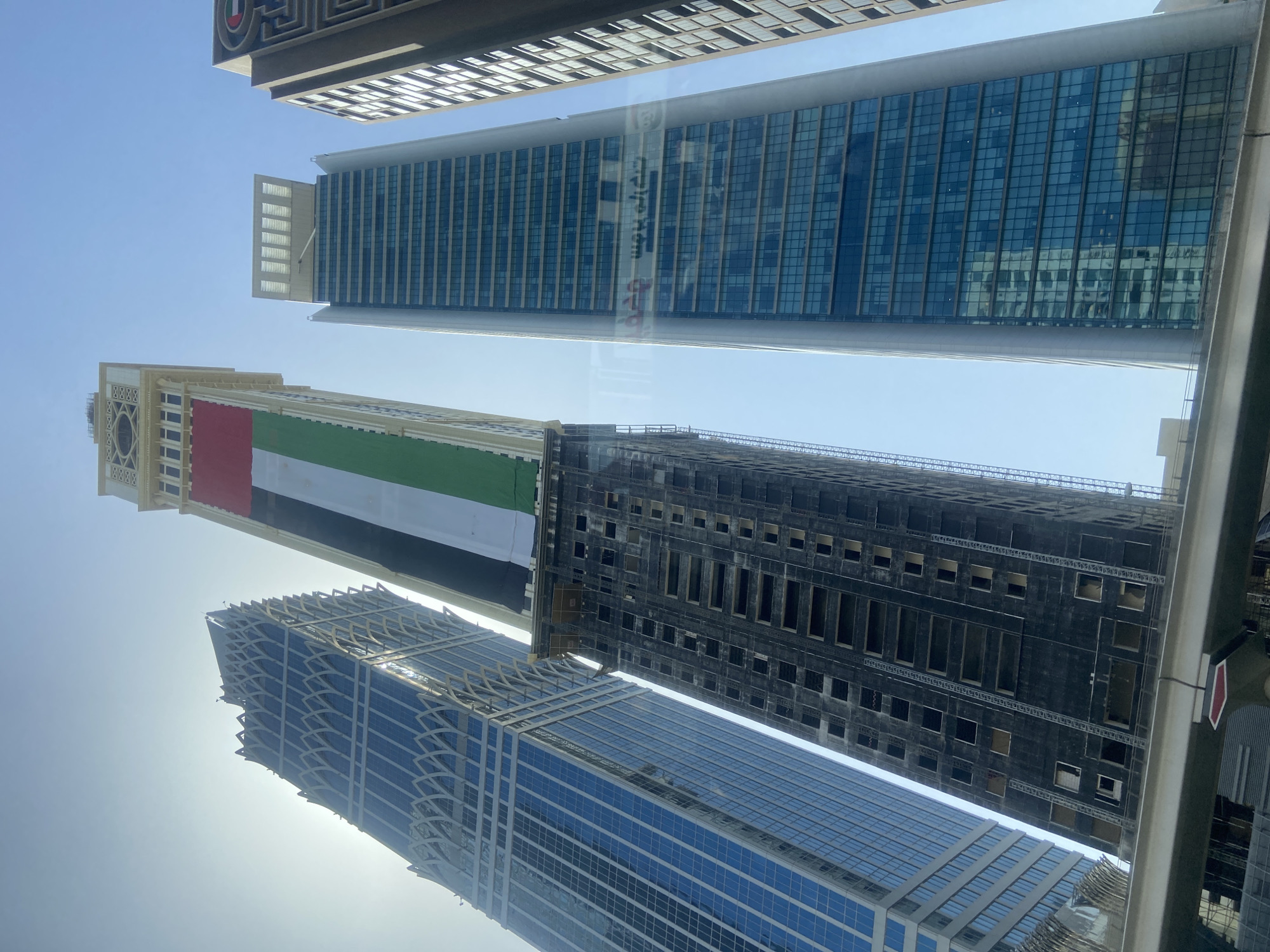 The image depicts a low-angle shot of several skyscrapers in Dubai, United Arab Emirates, against a clear blue sky. One of the buildings is decorated with the flag of the United Arab Emirates, featuring red, green, white, and black stripes. Another building appears to be under construction, with a dark, exposed facade. The other buildings have glass and steel facades.