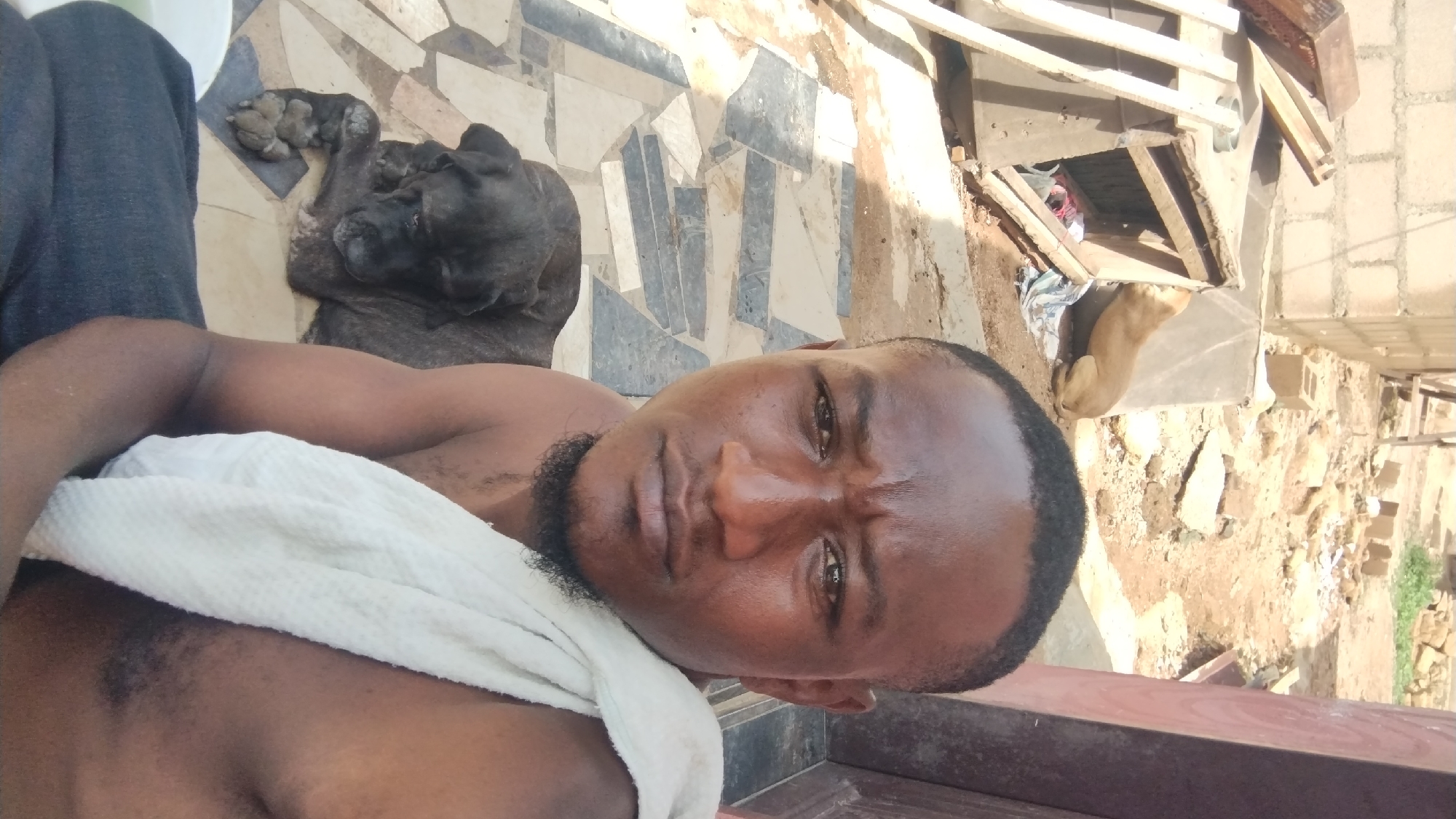 The image, taken in a selfie style, depicts a dark-skinned man resting outdoors, likely on a patio or porch. He lies on his back, looking directly at the camera with a neutral, slightly serious expression. He has a short beard and is shirtless, with a white, textured towel draped across his chest and left shoulder. His leg, covered in dark blue or grey pants, is partially visible on the left.
Beside the man, on a light-colored, irregularly patterned tiled floor, a large, dark-brindled dog is peacefully resting or sleeping. Its face is turned away, showing its dark fur and one paw. The tiled pattern consists of various grey and beige geometric shapes.
The background reveals an outdoor, somewhat rustic environment. It features loose dirt, scattered rocks, and debris, including wooden planks and what appear to be concrete blocks, suggesting a construction area or an undeveloped part of a property. Further back, a lighter-brown dog is visible, resting near some rubble and a partially obscured structure. The scene is brightly lit by natural sunlight, indicating daytime with clear weather. The overall atmosphere is casual and relaxed, set in what appears to be a domestic outdoor space in Jos, Nigeria. No visible text is present.