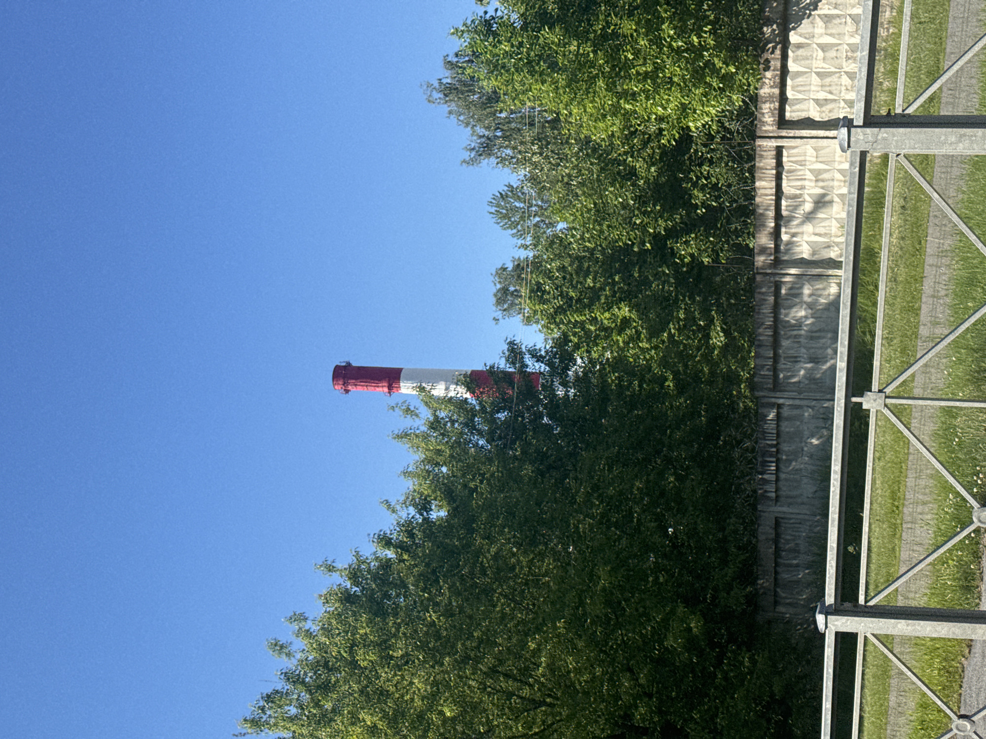 The image, oriented vertically, presents an outdoor scene dominated by a tall industrial chimney under a clear blue sky. The chimney, prominently featured in the upper left, is painted with alternating horizontal stripes of bright red and white.
A dense line of vibrant green trees or bushes extends horizontally across the middle section of the image, partially obscuring the base of the chimney and creating a lush green backdrop against the sky. The foliage appears healthy and full, suggesting a warm season.
In the foreground, a multi-part fence separates the viewer from the trees and chimney. To the left, a textured concrete wall, likely a precast fence panel, displays a geometric pattern of raised diamonds or squares, appearing somewhat weathered. To its right, and spanning the far right side of the frame, is a sturdy metal fence or railing made of light grey, possibly galvanized steel. This metal structure consists of vertical posts and diagonal cross-braces, with circular fastenings visible at intersections. Beyond this metal fence, a strip of green grass with scattered small white flowers is visible, suggesting a pathway or lawn area.
The sky is a uniform, bright blue, indicating clear weather. The sunlight is strong, casting shadows on the concrete fence, which suggests it is daytime, likely morning or afternoon. There are no people visible, and no discernible activity is taking place. The overall impression is a static landscape view, possibly of an industrial area bordering a green space. The style of the concrete fence is consistent with architectural elements found in the given location context of Russia. No text is visible in the image.
