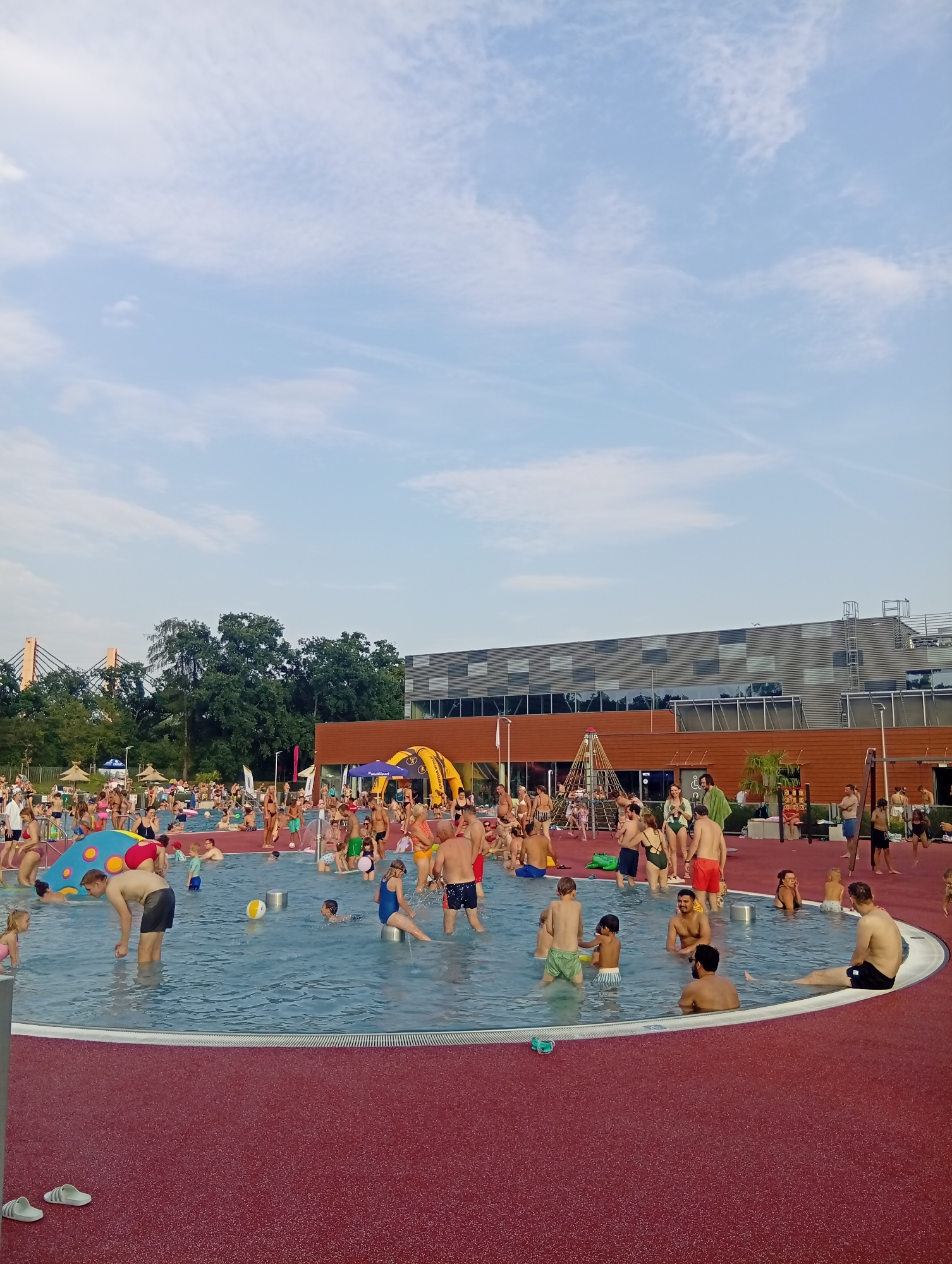 This is an exterior, eye-level, medium shot photograph of the Wrocław Aquapark, a large outdoor swimming pool complex in Wrocław, Poland, on a bright, cloudy day. The pool is filled with many people, including adults and children, who are swimming, standing, and interacting. The pool water is blue and the surrounding area is a reddish-brown color.
In the background, there is a large, modern building with a grey facade and a brown roof. There are also trees and a bridge visible in the distance. The sky is blue with some white clouds.
The people in the pool are wearing swimsuits and are engaged in various activities. Some are playing with balls, while others are simply relaxing in the water. The atmosphere appears to be lively and fun.
Specific details of note include:
* A large inflatable yellow structure in the middle ground, possibly a slide or play area.
* Several beach umbrellas providing shade.
* A children's play area with colorful structures.
* People sunbathing and socializing on the pool deck.
The image captures a typical summer day at a popular water park, with a focus on the recreational activities and the diverse crowd enjoying the facilities.