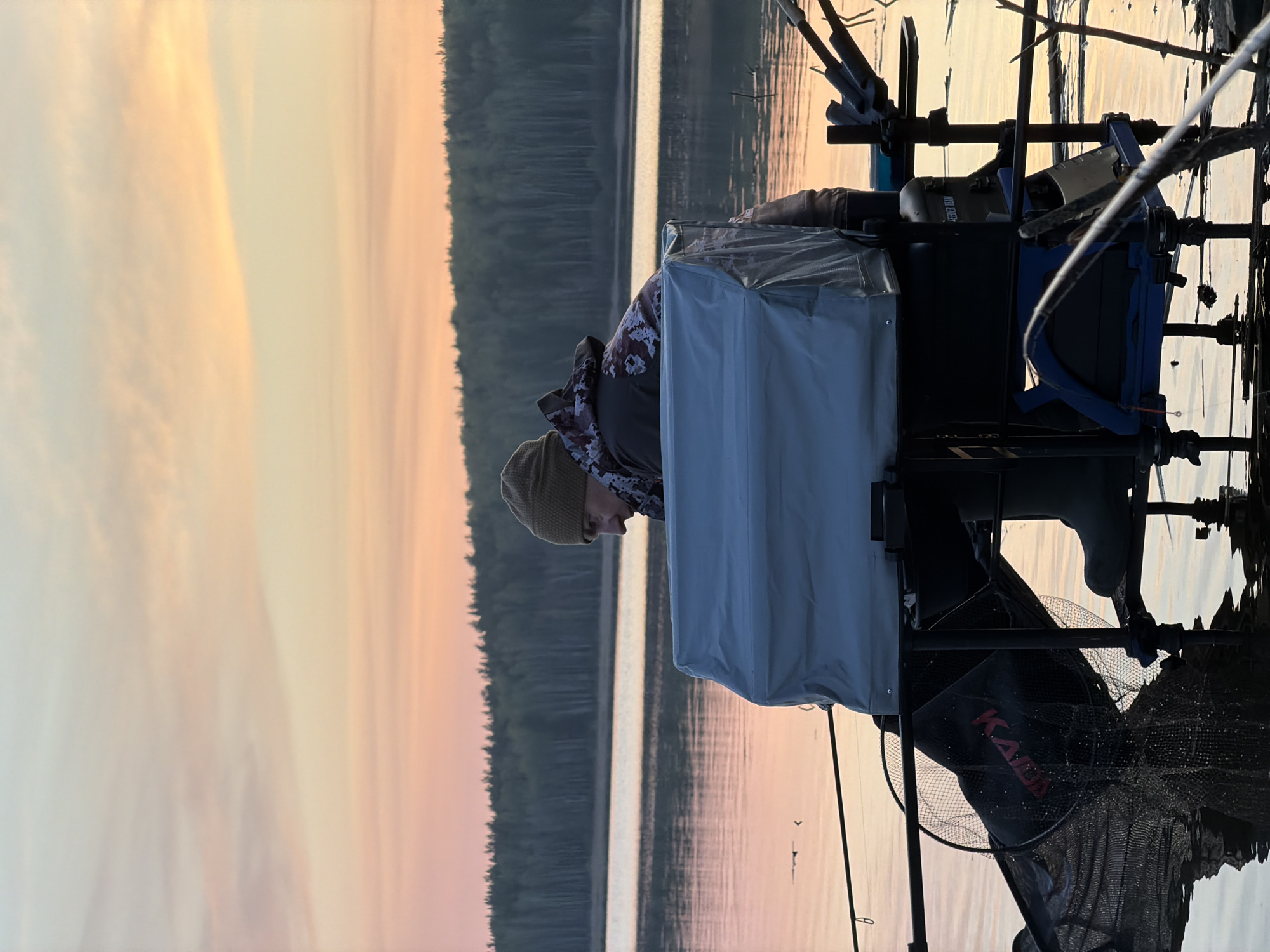 A person wearing a hat and camouflage jacket is fishing in a lake near Chaykovsky, Russia. The sun is setting, casting a golden light over the water and the forest in the background. The person is positioned next to fishing equipment including multiple fishing rods, nets and a square storage container.