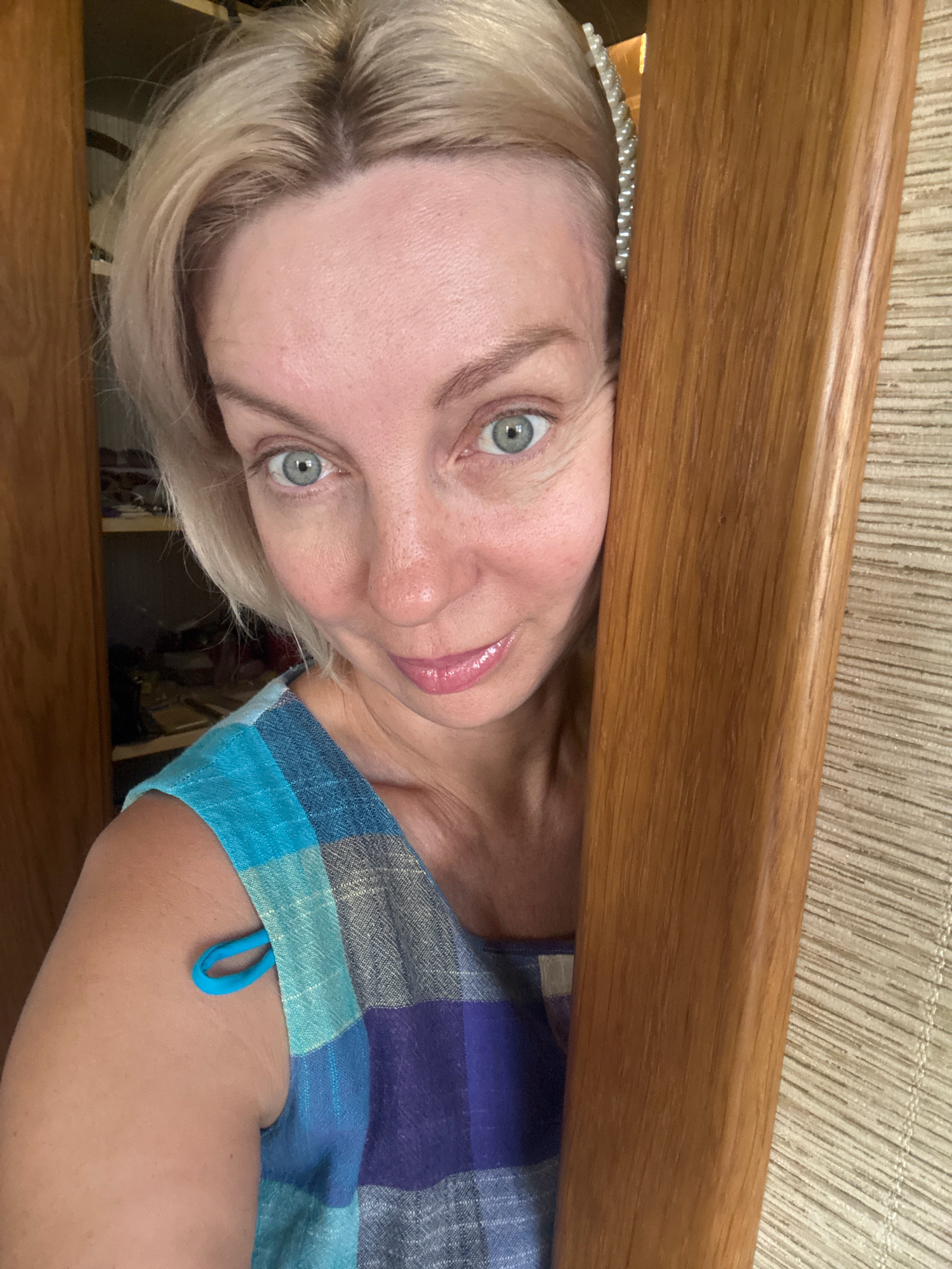 A blonde woman is peeking from behind a wooden door frame. She has blue eyes and is wearing a blue and purple plaid dress. There is a pearl hair clip in her hair. The background shows shelves with items and a textured wall. The scene is indoors.