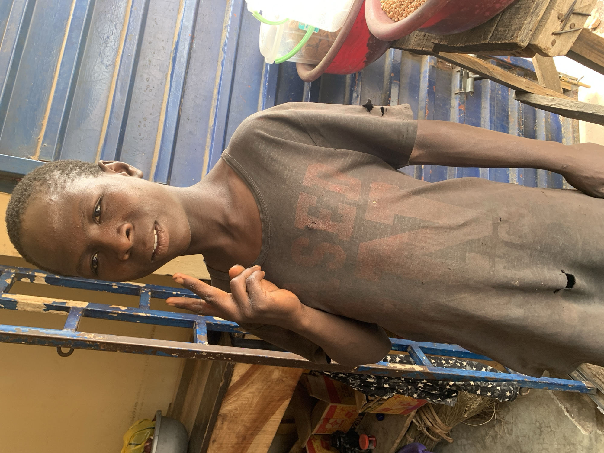 In a medium, slightly angled shot, a young, slender, dark-skinned person poses in Jos, Nigeria. The person is seen from the waist up, with the head tilted to the left. The individual is wearing a dark, worn t-shirt with a faded graphic on the front and several holes. They are making a peace sign with their right hand. The background includes a blue metal structure, a wooden surface with containers, and other miscellaneous objects below. The lighting is natural and seems bright.