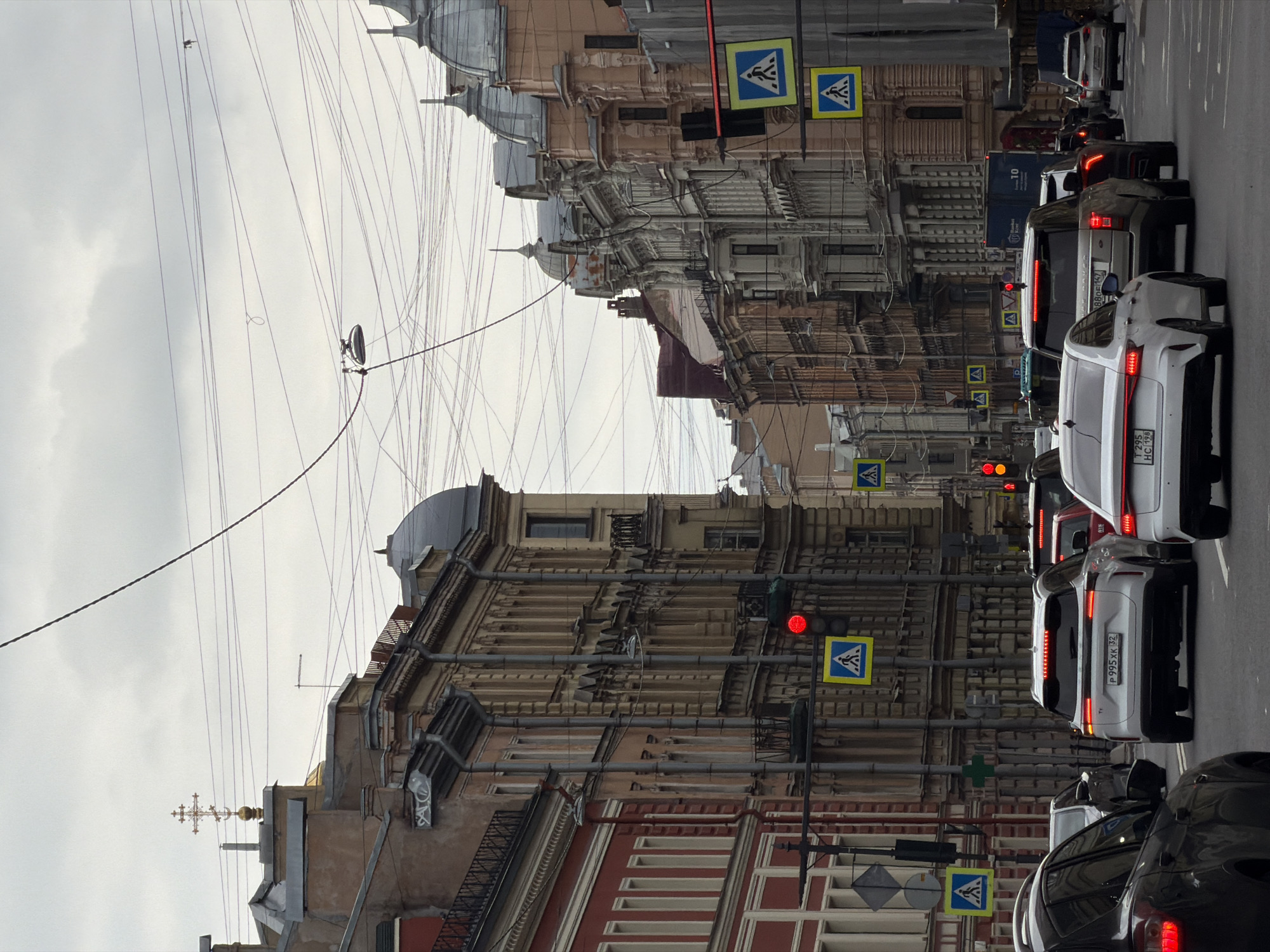 The image depicts a street scene in Saint Petersburg, Russia. The view is along a street lined with old, ornate buildings, showcasing detailed architectural elements. Overlapping the buildings are multiple overhead cables that crisscross the sky. The street itself is congested with vehicles, mostly cars, stopped at red traffic lights. There are numerous pedestrian crossing signs along the street, indicating pedestrian pathways. The sky overhead is overcast and gray. The photo captures a mix of historical architecture and modern urban congestion.