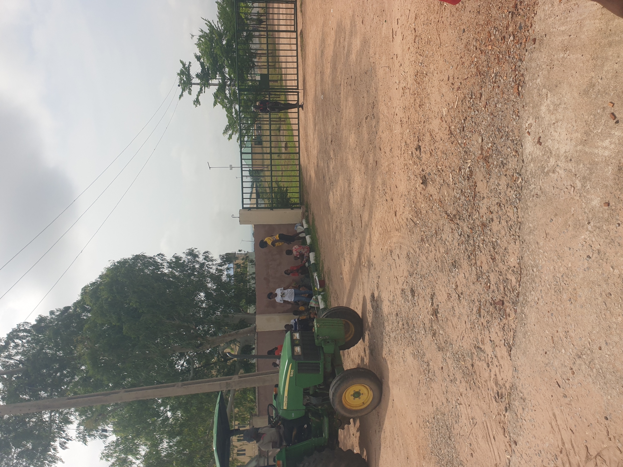 In Jalingo, Nigeria, a green John Deere tractor with a man operating it is positioned on a dirt road. To the right of the tractor, a group of people, including children, are gathered near a beige wall and a tall metal gate. One person, wearing a yellow shirt, stands near the gate. The environment appears to be an outdoor setting with trees and a cloudy sky. The ground is covered in dirt and small stones, and there are some scattered pieces of debris. The overall scene suggests a community gathering or activity taking place outdoors in a semi-urban environment.