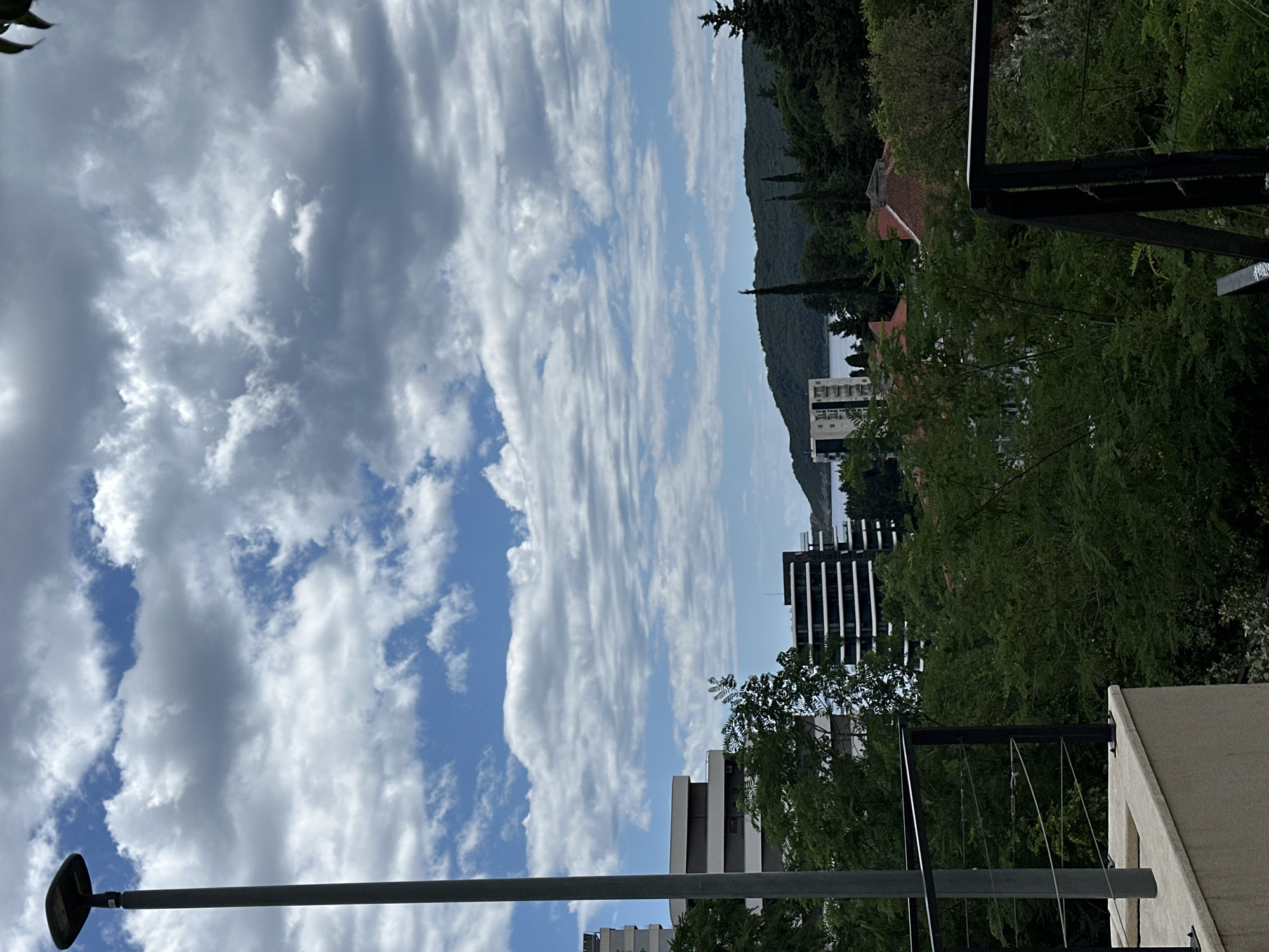A street lamp pole stands to the left of the image, with a gray pole and black lamp. In the background, there are several modern buildings with dark facades and white balconies, nestled among lush green trees. A body of water is visible in the distance, with a green, tree-covered hill forming the horizon. The sky is filled with a mix of white and gray clouds against a blue backdrop. The foreground shows a portion of a concrete balcony with a metal railing. The scene appears to be a view from a higher vantage point, possibly a residential building or a hotel in Bečići, Montenegro, given the architectural style and the coastal landscape.