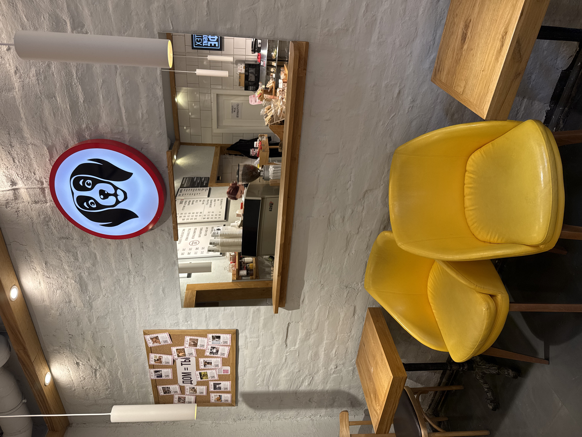 The image depicts the interior of a café or restaurant, likely in Saint Petersburg, Russia, as suggested by the Cyrillic text "ДОМ = ТЫ" (HOME = YOU) on a bulletin board. The wall is painted white with a textured finish. A bright, round sign with a stylized dog face is mounted on the wall. Above and slightly to the right of the sign is a large mirror reflecting the café's counter area, showcasing pastries, coffee cups, and staff. Below the sign is a cork bulletin board with various pictures and notes. Two cylindrical lights hang from the ceiling. The foreground features a stack of two bright yellow chairs next to a wooden table. The scene is well-lit. There are no visible people interacting in the immediate frame.