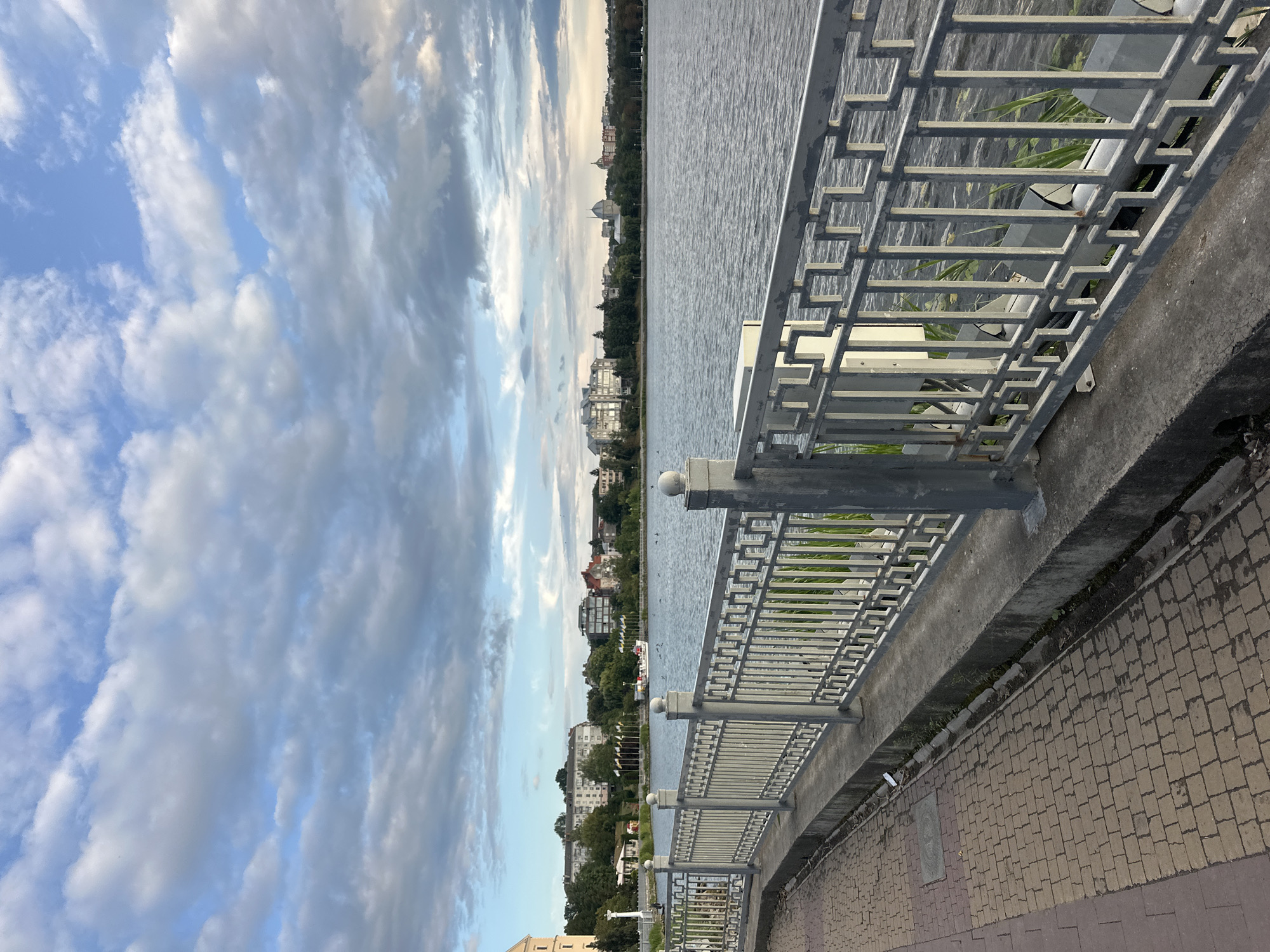 The image depicts a view of Ternopil Lake in Ternopil, Ukraine. In the foreground, a cobblestone sidewalk curves along the edge of the lake, bordered by a decorative metal railing. Beyond the railing, the lake stretches out, with a boat visible in the distance. The opposite shore is lined with trees and buildings, including some apartment complexes and what appears to be a church with a dome. The sky is partly cloudy, with a mix of blue sky and white and gray clouds. The overall scene suggests a pleasant, perhaps late afternoon or early evening atmosphere.