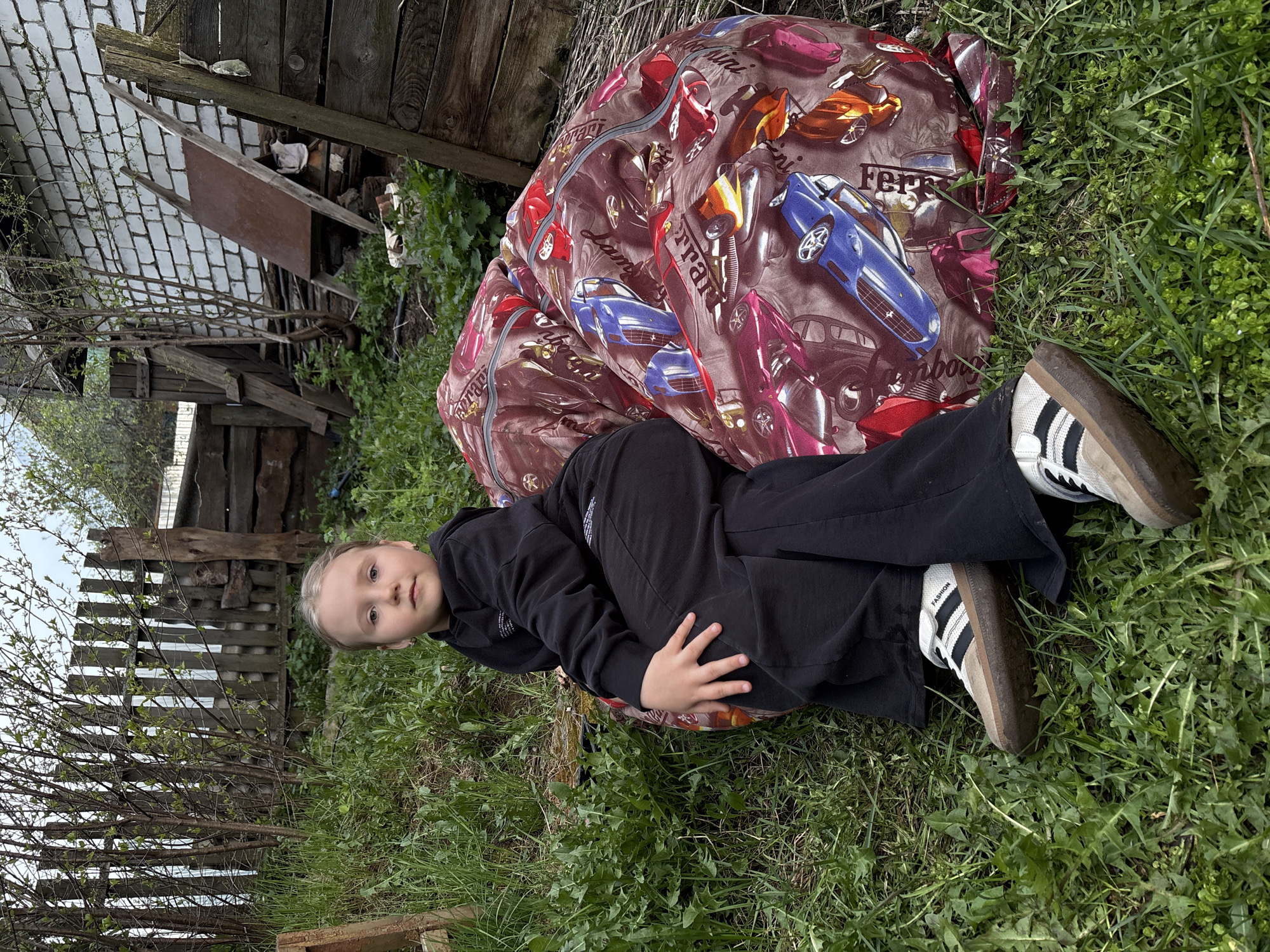 A young child is lying on a brown, car-themed beanbag in a grassy area. The child is wearing a black hoodie and black pants, and white sneakers with black stripes. The location appears to be an outdoor area, possibly a backyard, in the city of Chaikovsky, Russia. In the background, there is a wooden fence and a brick structure. The sky is visible in the upper left corner, suggesting it is daytime.