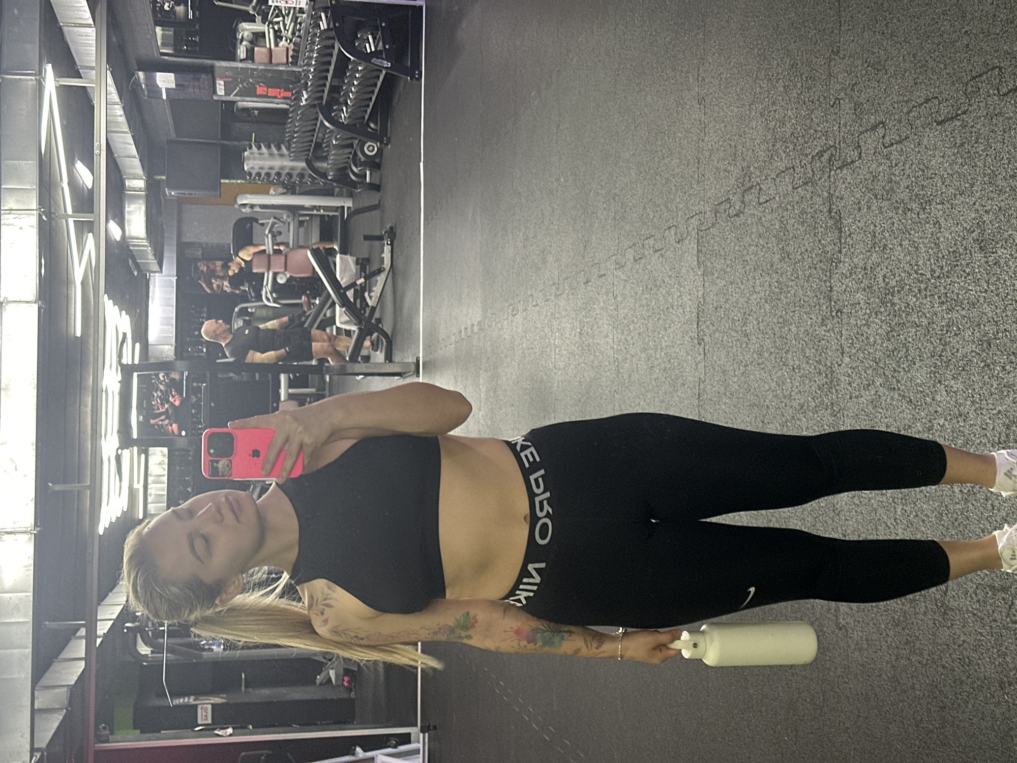 The image depicts an indoor gym setting, featuring a woman taking a self-portrait with her phone. The main subject, standing in the foreground, is a woman with long blonde hair tied in a ponytail. She is wearing a black sports bra and black leggings that prominently display the text "NIKE PRO" on the waistband, with "NIKE" and a partially inverted "PRO" visible. Her left arm is adorned with extensive floral-themed tattoos. She holds a bright pink smartphone in her right hand, pointed towards herself, and a large white water bottle in her left hand. Her expression is neutral, with her eyes slightly narrowed.
In the background, several other individuals are visible, actively engaged in workouts on various gym equipment. One bald man wearing dark attire is situated in the mid-ground, utilizing a weight machine. The gym is equipped with rows of dumbbells, various weight machines, and benches. The flooring consists of dark gray interlocking rubber mats. Overhead, exposed metal ductwork and artificial lighting fixtures are visible. The overall ambiance suggests a functional workout environment. No specific location cues for Budva, Montenegro are discernible within the image itself.