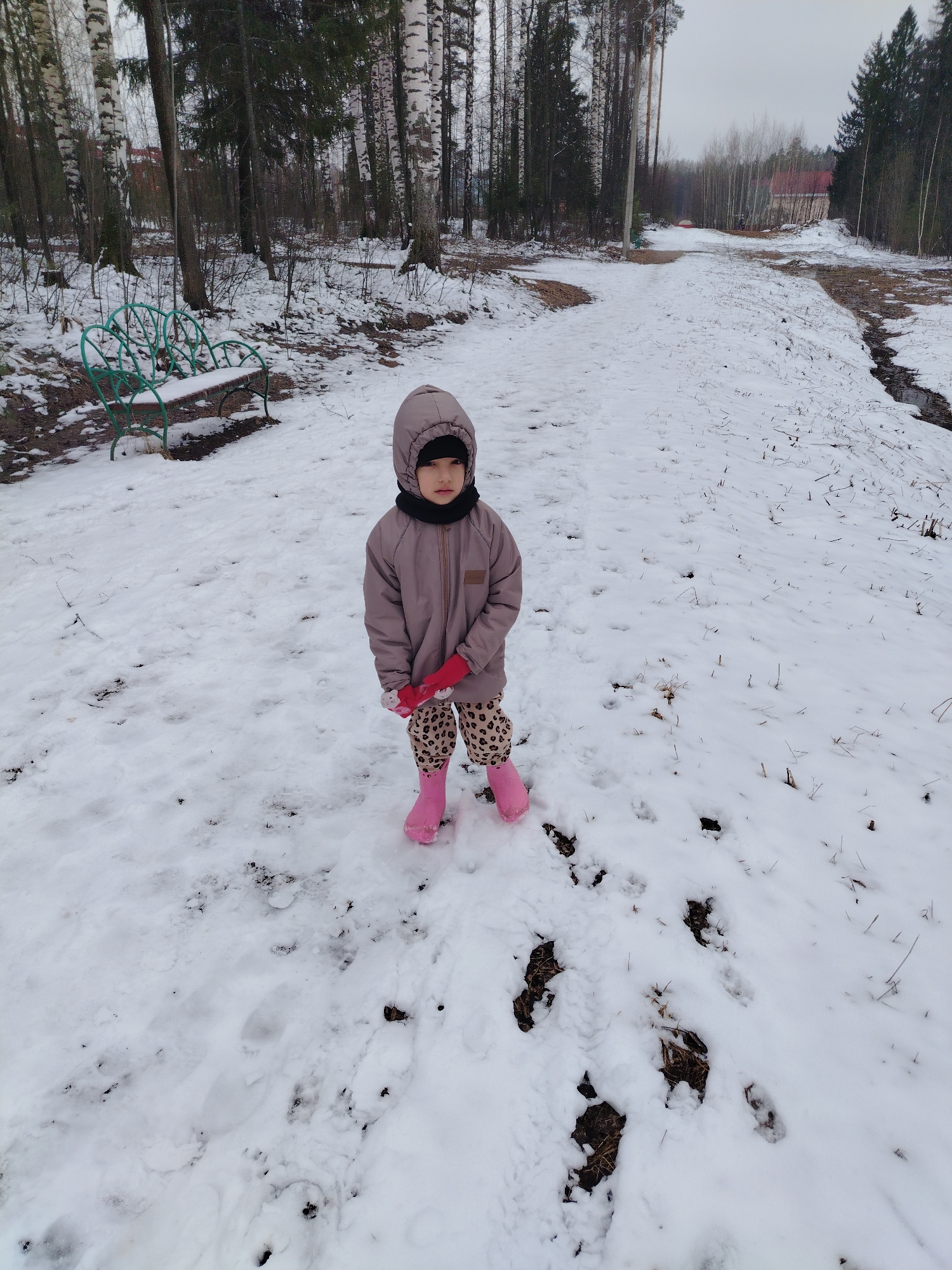 A young child stands in a snowy landscape, likely in Noviy, Russia, given the prompt. The child wears a hooded jacket, patterned leggings, pink boots, and gloves. Bare trees and a sparse forest line the background. A green bench sits to the left of the child. A dirt path covered in snow extends into the distance. The ground is covered in patches of snow and exposed earth. The sky appears overcast.