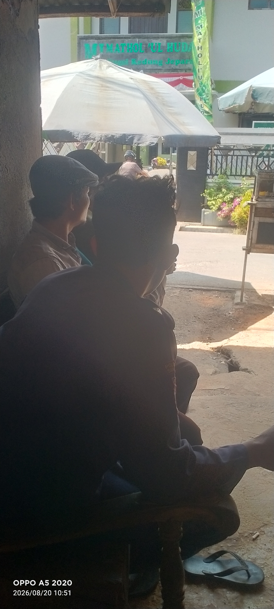 In Bugel, Indonesia, two men are seated outdoors. The man on the left wears a cap and a light-colored shirt, and the man on the right is in the foreground, facing away from the camera, wearing a dark shirt. Behind them, a white umbrella is open, and a building with signage that reads "MT MATHOL 'UL HUDA" is visible. A black metal fence separates the area from a street with some foliage and a small market stall. The ground is a mix of dirt and concrete, with a discarded flip-flop visible near the seated men. The lighting suggests it is daytime, with shadows cast on the ground.