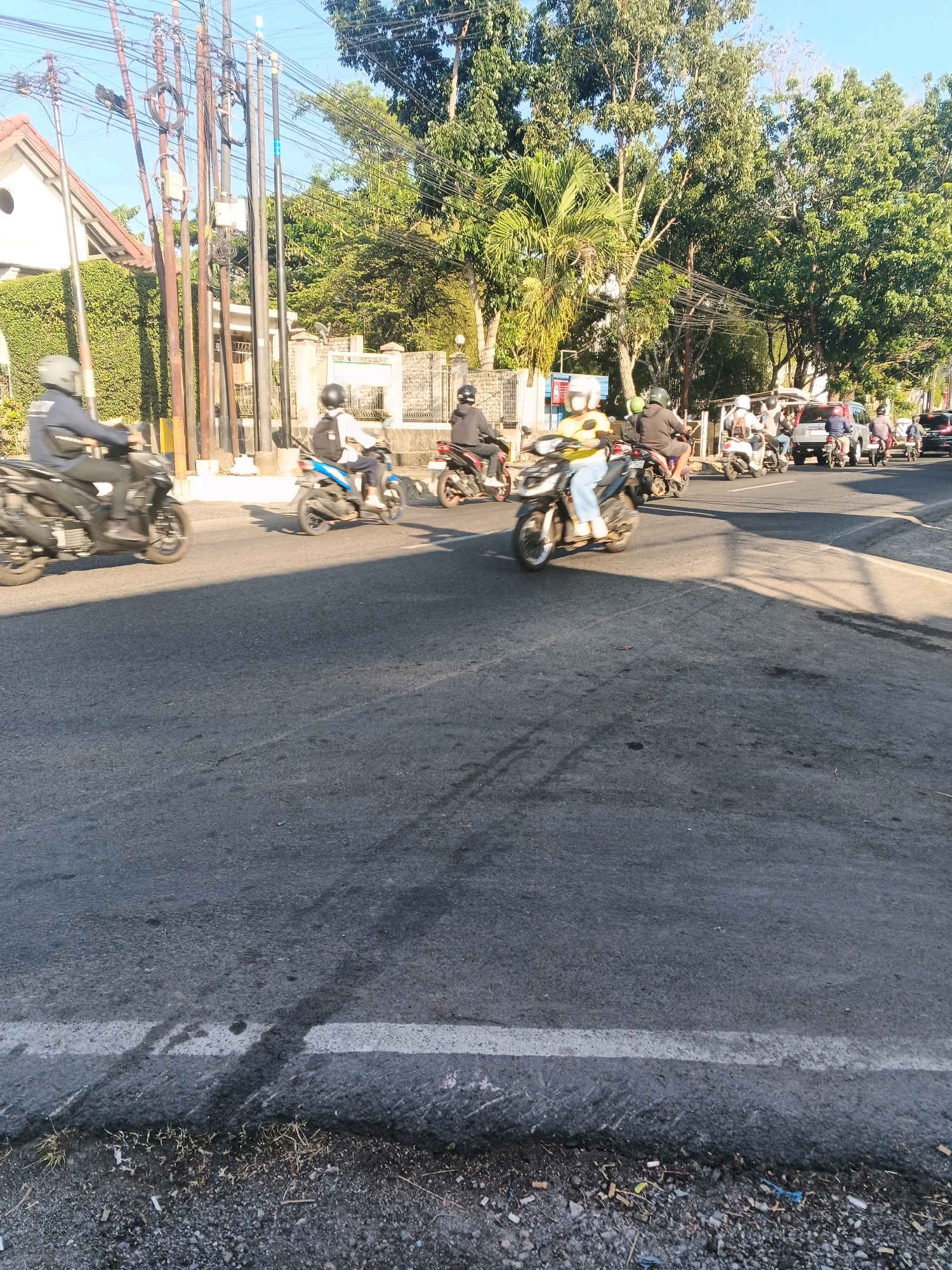 The image depicts a street scene, likely in Semarang, Indonesia, given the visual cues such as the type of buildings and vegetation. The focus is on a road with several motorbikes and a few cars. The weather appears to be sunny with clear skies, and the light suggests it's either morning or late afternoon.
In the foreground, a dark asphalt road dominates the lower half of the frame, marked by a white line at the edge. The road surface shows some wear and tear, with patches of darker discoloration and debris on the side.
Mid-ground, the street is active with vehicular traffic. Multiple individuals on motorbikes are clearly visible, most wearing helmets for protection. One person in the foreground is on a dark motorbike, wearing a dark jacket and helmet, moving from left to right. To their right, another motorbike is ridden by a person in a white shirt and helmet, with a backpack. Further back, more motorbikes and a few cars are in motion. One car, a silver SUV, is prominent among the vehicles.
The background features a mix of urban and natural elements. A house with a tiled roof and white walls is partially visible on the left. Numerous utility poles with tangled wires are erected along the roadside. Lush green trees, including palms, provide a dense canopy. A stone wall with an archway suggests the entrance to a property or institution. The overall impression is a busy urban road bordering a more residential or verdant area.