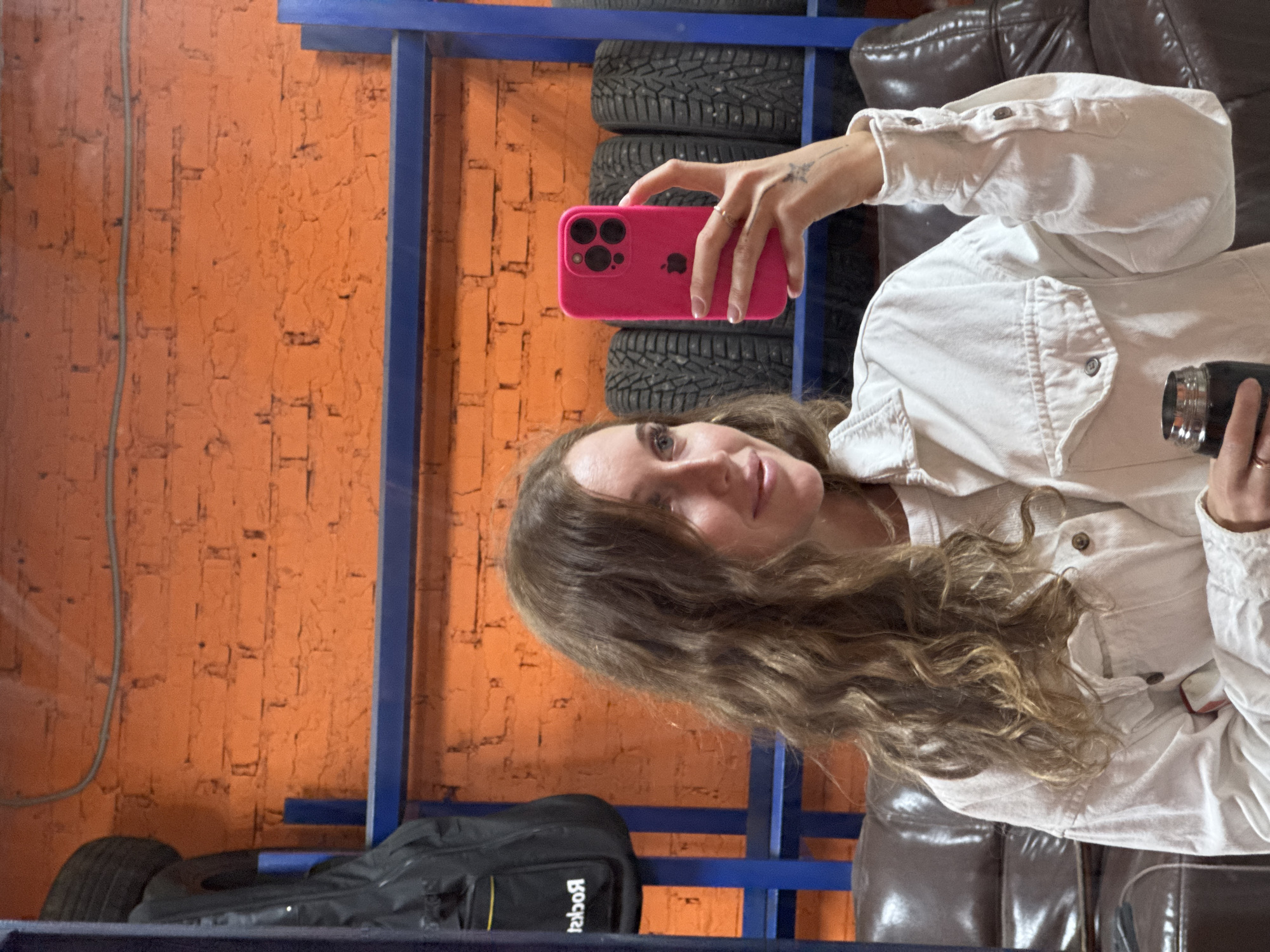 A woman takes a selfie with a pink iPhone in a room with a stack of tires on a blue metal rack and an orange brick wall. A black duffel bag rests on the bottom rack. She wears a white denim jacket and sits on a brown leather couch. The location is Chaikovsky, Russia.