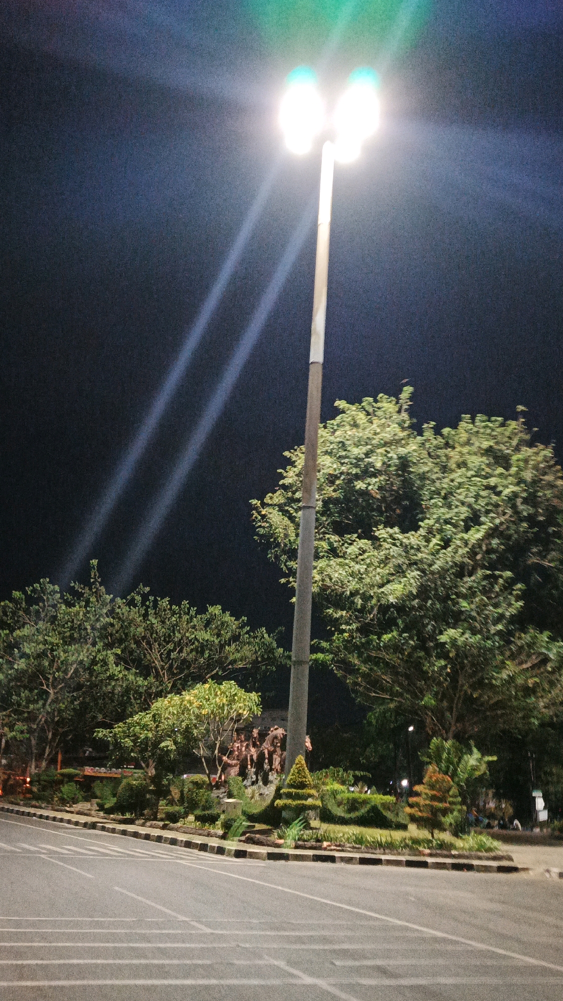 This image captures a night scene at a roundabout in Surakarta, Indonesia, with a prominent dual-headed streetlamp illuminating the area. The streetlamp, positioned on a tall pole, emits bright, cool-toned light, casting distinct beams across the dark sky.
The roundabout itself is paved and marked with white lane lines, indicating it's an active traffic area. Surrounding the roundabout is a well-maintained garden area featuring various types of trees and meticulously sculpted bushes. A notable sculpture or monument, appearing to depict figures, is situated within this garden.
The background is predominantly dark, with the night sky serving as a backdrop to the illuminated streetlamp and the green foliage of the trees. The overall impression is a well-lit urban landscape at night, emphasizing the functionality of the streetlamp in enhancing visibility and the aesthetic appeal of the landscaped roundabout.