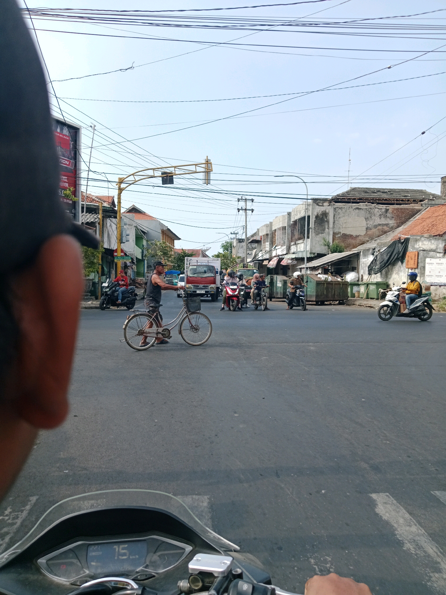 This is a street-level, medium shot from the perspective of someone riding a motorcycle, looking forward and slightly to the left. The scene depicts a busy intersection in Probolinggo, Indonesia. In the foreground, the handlebars and dashboard of a motorcycle are visible, showing a speed of 15 km/h. A person's hand is resting on the right handlebar.
The intersection is paved with asphalt and has white lane markings. Several vehicles are present: a man is walking and pushing a bicycle with a basket on the front, a red truck is stopped at the intersection, and multiple motorcycles with riders are also present. Some of the motorcycle riders are wearing helmets. In the background, there are buildings with varying states of repair, some appearing older and others under construction or renovation. Overhead, a network of electrical wires crisscrosses the sky, typical of urban areas in Indonesia. A traffic light pole with signal lights is visible in the mid-ground. The sky is clear and bright, suggesting a daytime scene. A billboard with red and white text is partially visible on the left side of the image. The overall atmosphere is one of everyday urban life and traffic.