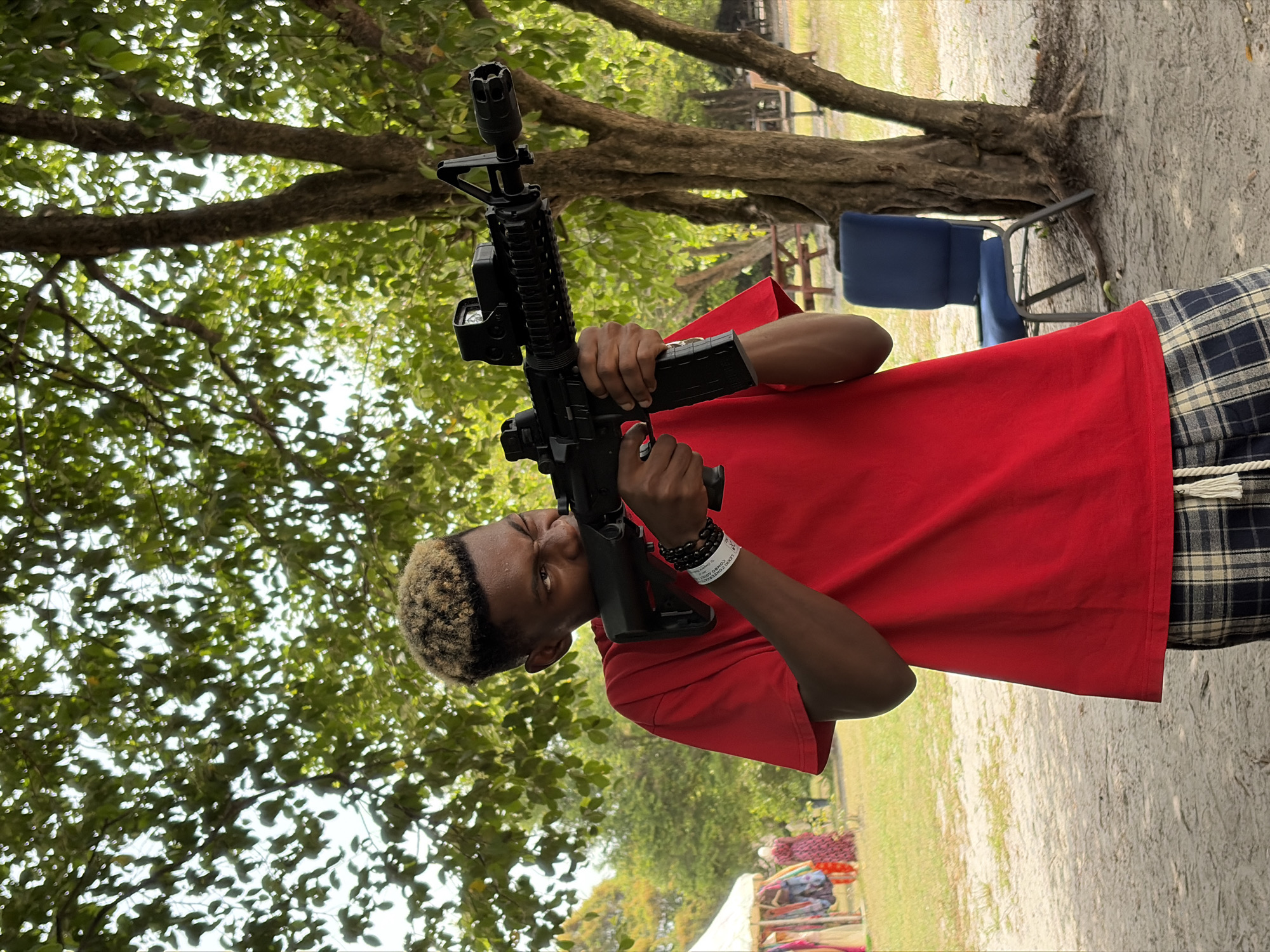 An individual is holding a black AR-15 style rifle in a slightly wooded area. The person is wearing a red t-shirt and plaid pants. There is a blue chair in the background near a tree. The location is possibly in Lekki, Nigeria. The individual is aiming the rifle upwards. There are trees overhead. There appear to be items for sale in the background on the lower-left. The individual is wearing a bracelet and a wristband.