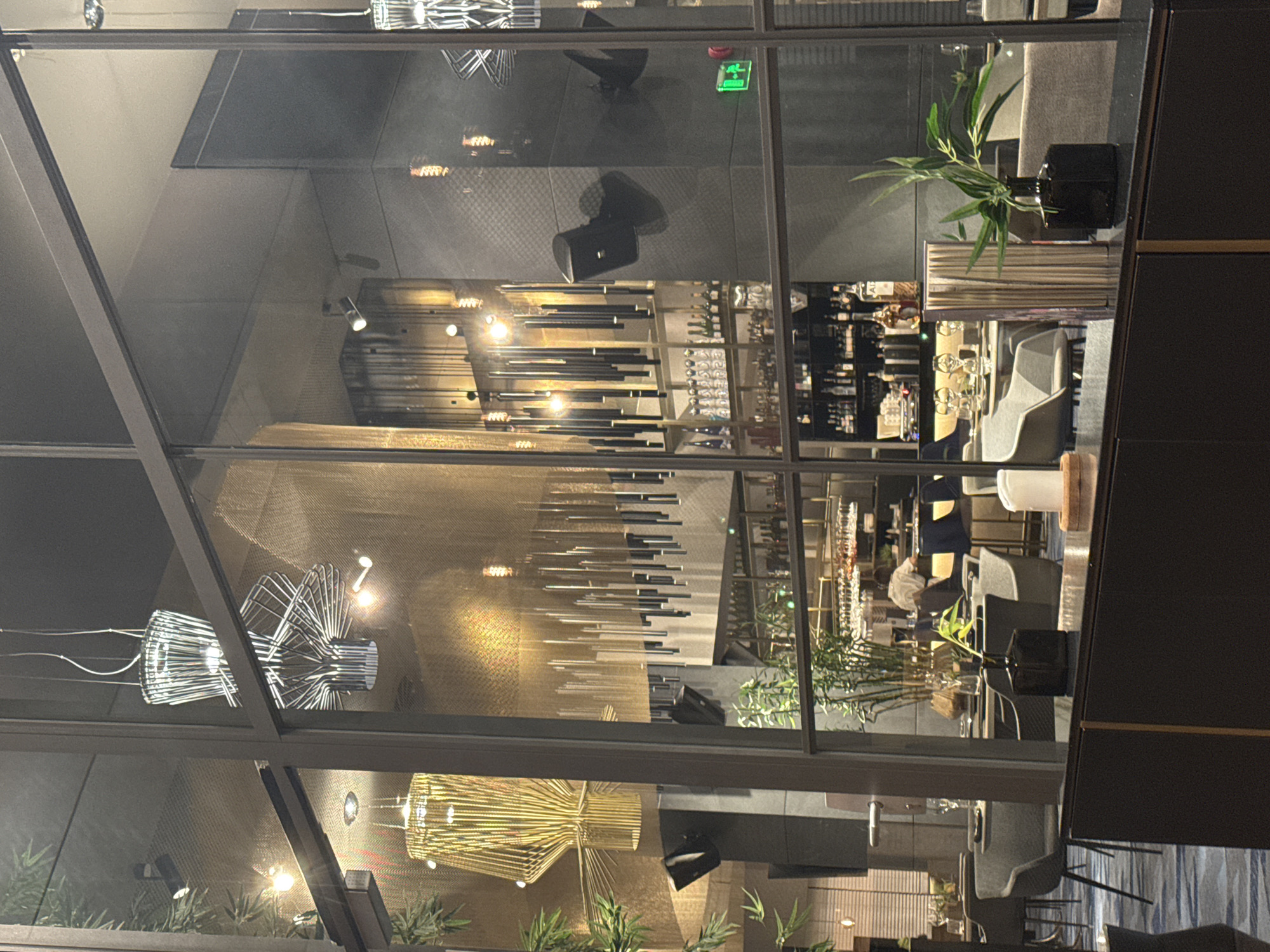 The image shows the interior of a restaurant, likely located in Sofia, Bulgaria, as evidenced by the "EXIT" sign in Cyrillic. The restaurant appears to be modern and upscale, with large windows reflecting the interior.
Inside, diners are seated at tables set with white tablecloths and place settings. The tables are spaced apart, and the lighting is dim and ambient, with several decorative chandeliers and pendant lights visible. In the background, a bar area is stocked with bottles of alcohol, and glassware is neatly arranged on shelves. A server can be seen behind the bar, wearing a white shirt.
The foreground of the image features a dark countertop with decorative items, including a vase with greenery and a white candle on a wooden holder. The overall ambiance suggests a sophisticated dining experience.
