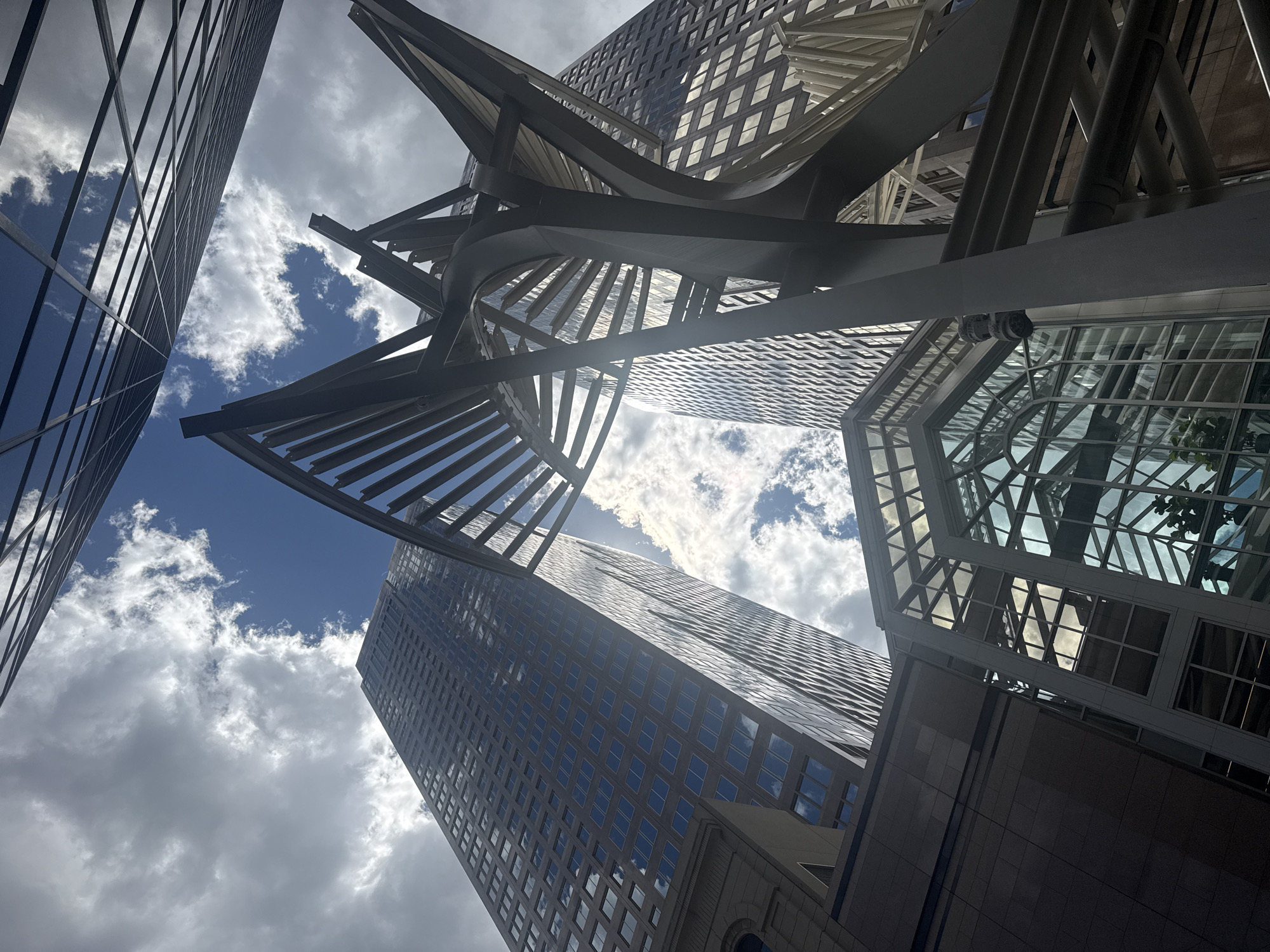 This is a low-angle shot of the Bow Building in Calgary, Canada, on a cloudy day. The sky is visible through the building's modern architecture, with patches of blue sky and white clouds. The Bow Building, a distinctive skyscraper, dominates the scene. In the foreground, a contemporary metal sculpture with a skeletal design and crisscrossing beams is positioned. Below the sculpture, a glass atrium with angular lines and reflective surfaces is visible. The overall scene captures the urban landscape with a focus on architectural elements and the sky.
