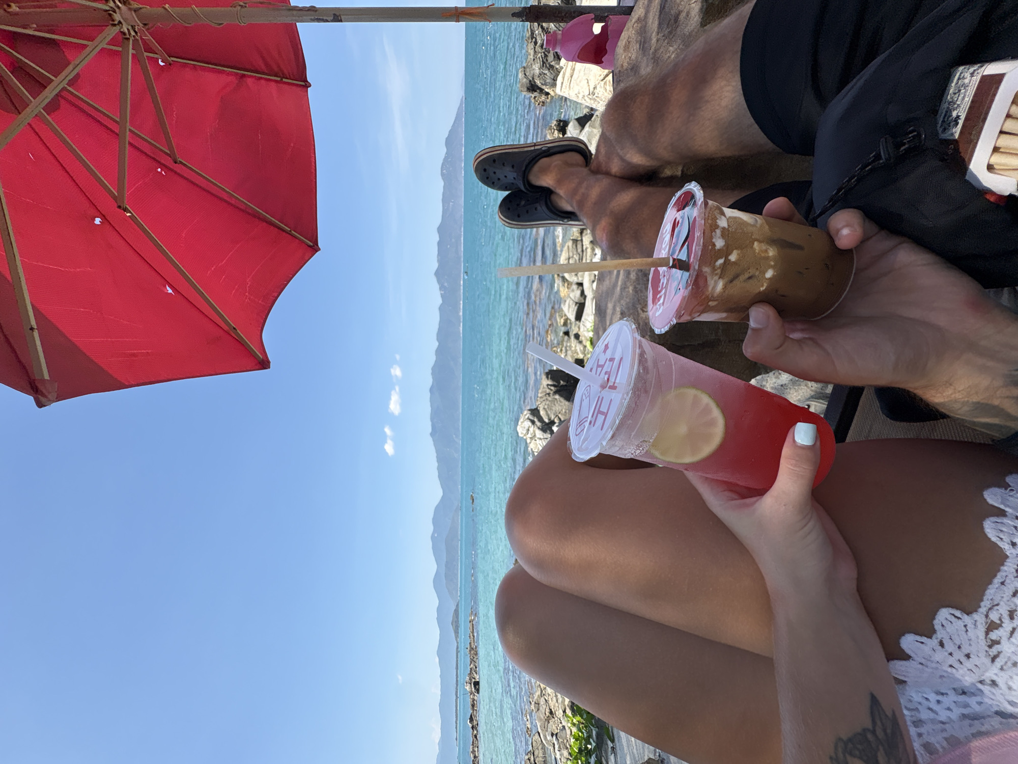 The image shows a view of a tropical beach scene, likely near Vĩnh Lương, Vietnam given the mountainous terrain in the background over the ocean. Two individuals are present, each holding a beverage in a plastic cup with straws. One drink is a red liquid with a lime slice, held by someone with fair skin and a small tattoo on their arm. The other drink is a brown coffee drink. The individuals appear to be sitting under a red umbrella for shade. The scene includes a turquoise ocean, rocky shoreline, and a clear blue sky. One person is wearing black shorts and black Crocs-style sandals.