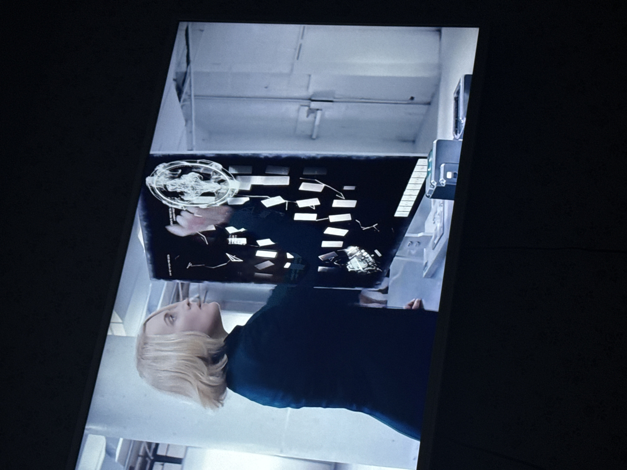 The image is a vertically oriented shot of a screen, likely a television or monitor, showing a scene from a film or television show. The angle is slightly off, suggesting the screen was photographed rather than a direct capture. A blonde woman in a dark garment is visible on the screen, looking up at a computer display filled with data, including what appears to be a circular graphic and multiple rectangular items, likely representing digital files. The background on the screen includes an industrial-looking ceiling and equipment, suggesting a technologically advanced setting. The image is set against a stark black background, further emphasizing the screen as the central element. No evidence indicates this is in Mytishchi, Russia.