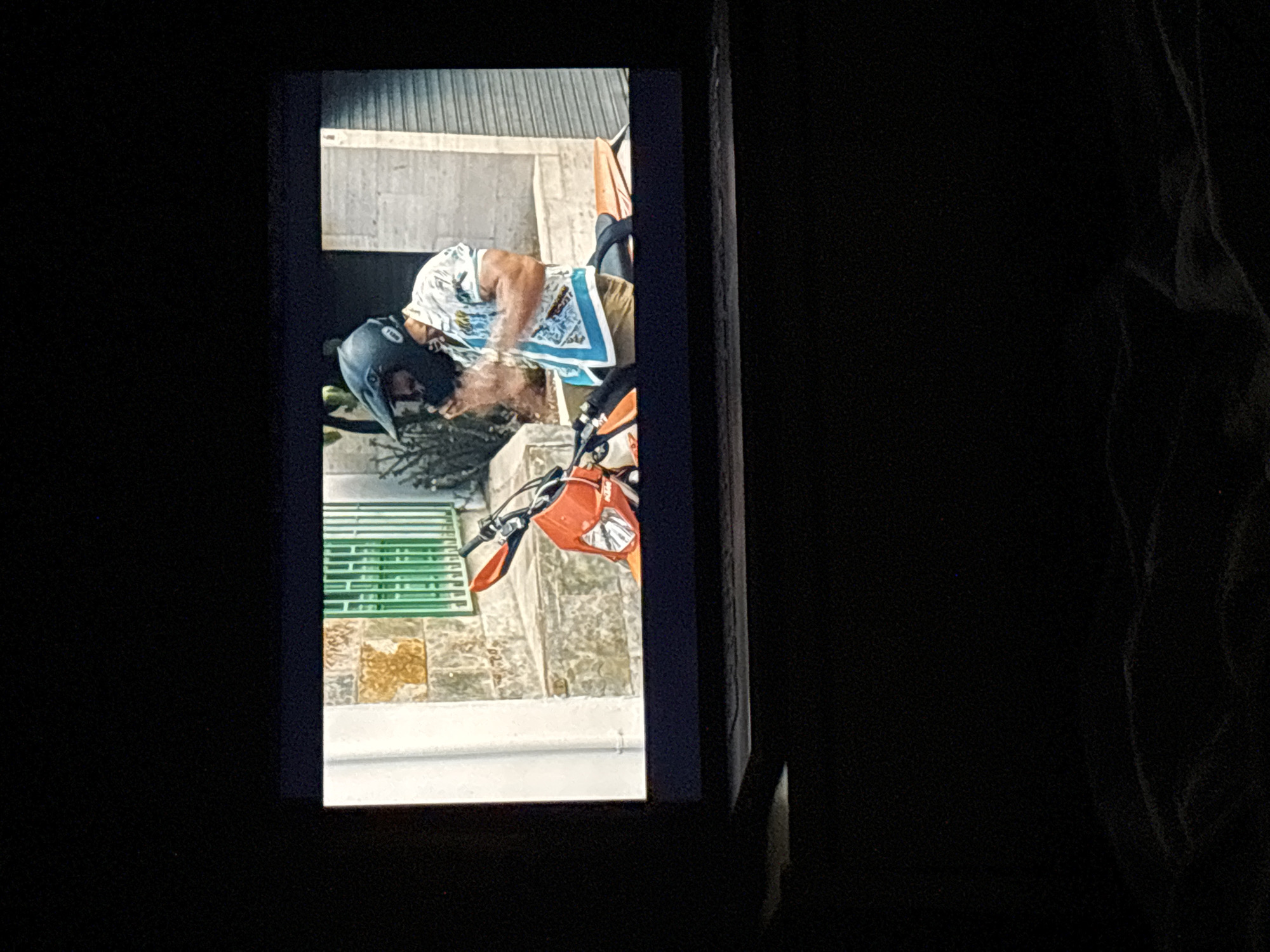 A digital image is displayed on a screen, viewed from a slightly elevated perspective in a darkened environment. The bottom foreground shows dark, textured fabric, likely a blanket or bedding.
The screen content depicts an outdoor scene in what appears to be a narrow street or passage within the city of Budva, Montenegro. A male individual, positioned centrally, is seated on or standing next to an orange dirt bike. He wears a black full-face motorcycle helmet with a prominent visor, displaying a white "BELL" logo on the chin bar, and has a visible beard. His attire consists of a short-sleeved, light-colored collared shirt featuring blue patterned motifs and a distinct blue border along the bottom and sleeve cuffs. His hands are raised in front of him, possibly adjusting the helmet or performing an action.
The motorcycle is an orange dirt bike, with the front fairing, headlight, and handlebars with orange handguards clearly visible. A faint "KTM" logo is discernible on the side panel. The seat is black.
The background consists of stone architecture. To the left, a wall constructed from irregular, light-colored stone blocks exhibits patches of yellowish discoloration, with a vertical white drainpipe running down its surface. A green metal grate or window barrier with vertical slats is positioned behind the individual. Further right, a building with a light-colored facade and a closed, dark corrugated metal roll-down shutter is visible. Sparse green vegetation is present in the background. The ground is partially visible and appears to be paved or cobblestoned.